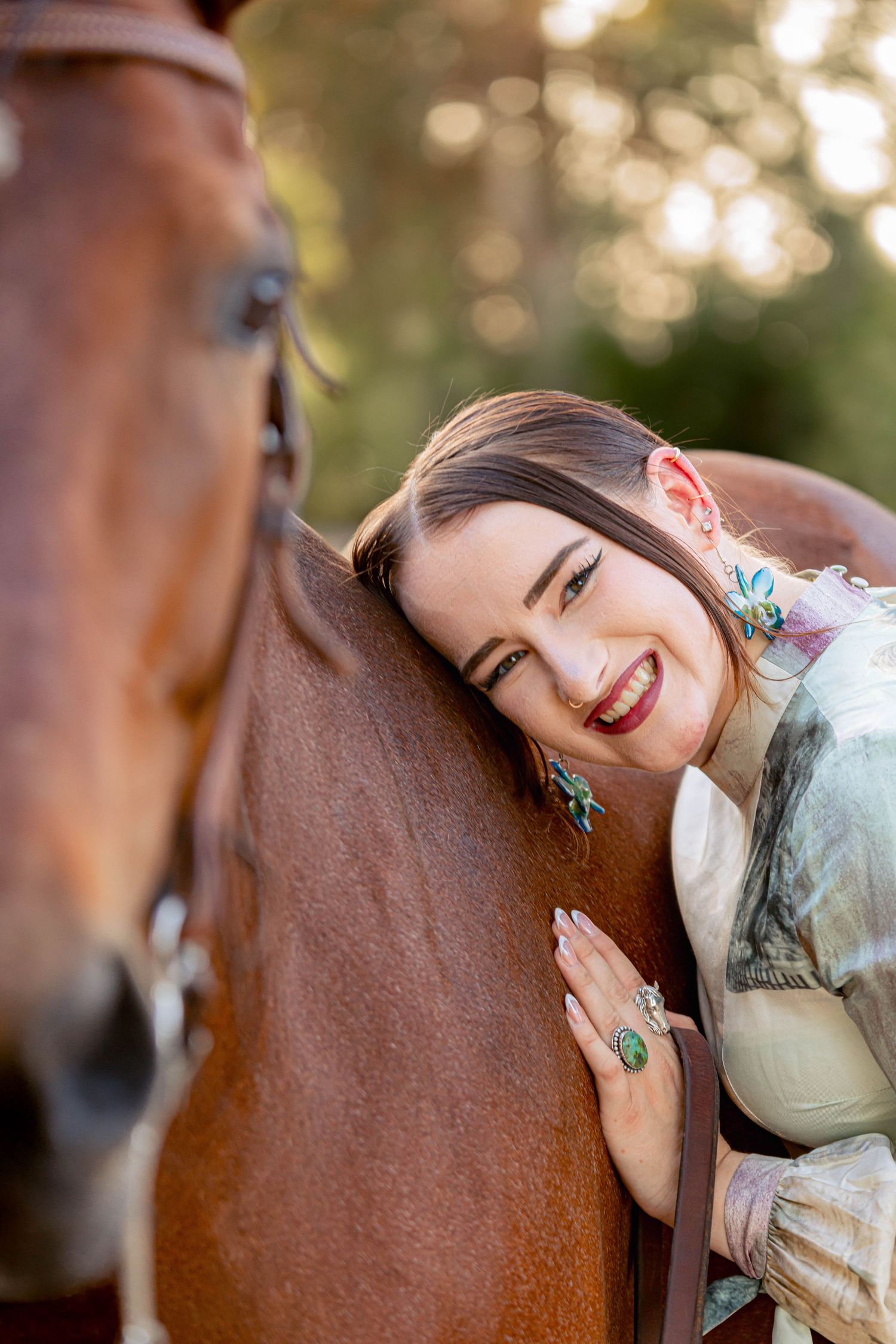 Smiling young woman in floral dress leaning against a chestnut horse outdoors in golden light.