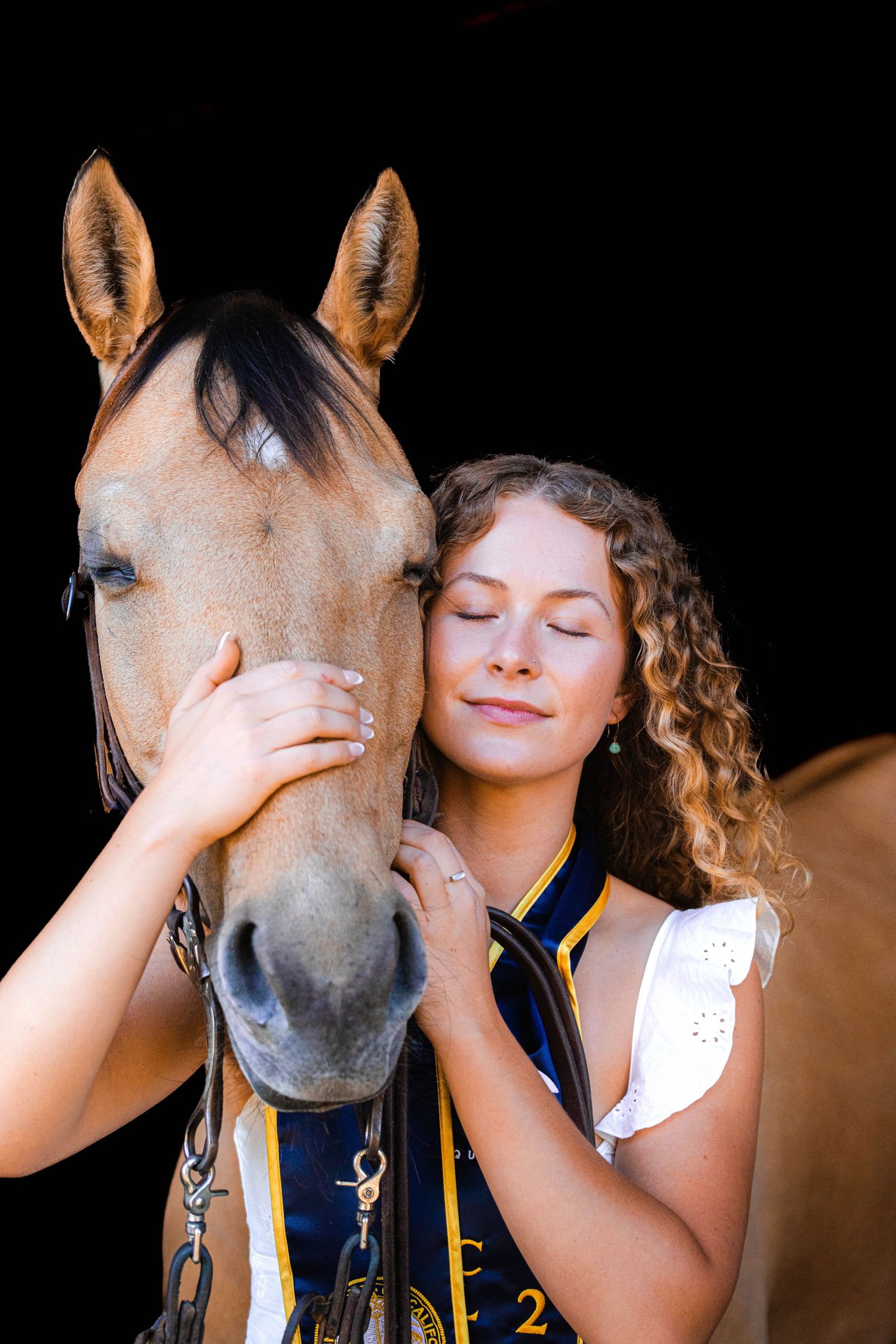 Curly-haired girl in varsity jacket closes eyes while gently hugging palomino horse face.