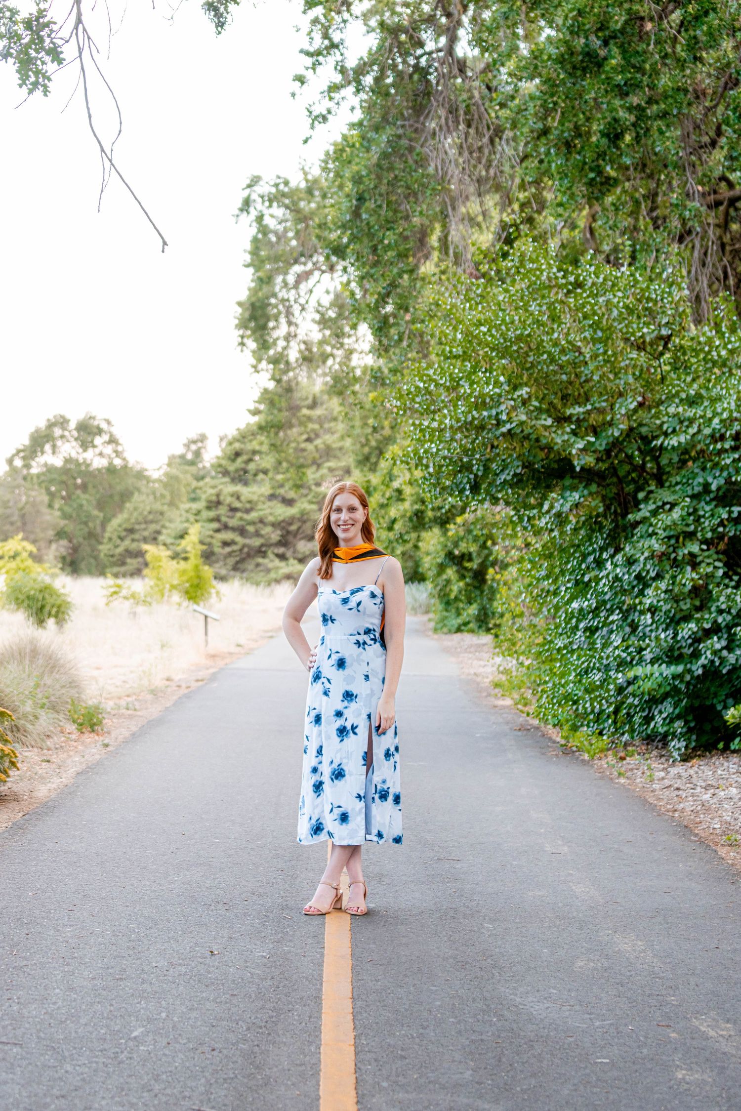 Smiling redhead in blue floral sundress standing on center yellow line of tree-lined road in bright daylight.