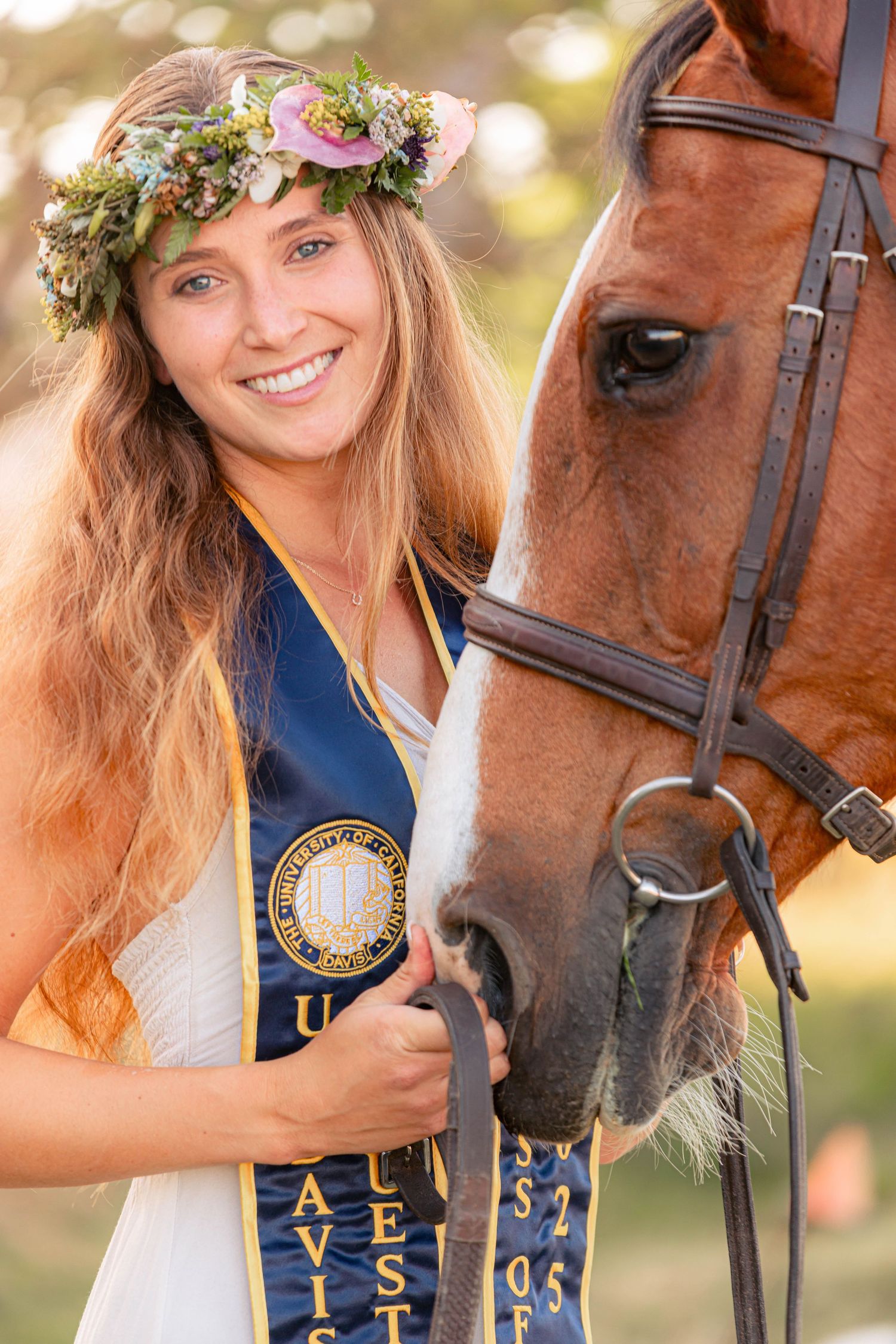 Smiling graduate wearing floral crown and UC Davis sash poses with chestnut horse in sunny outdoor setting.