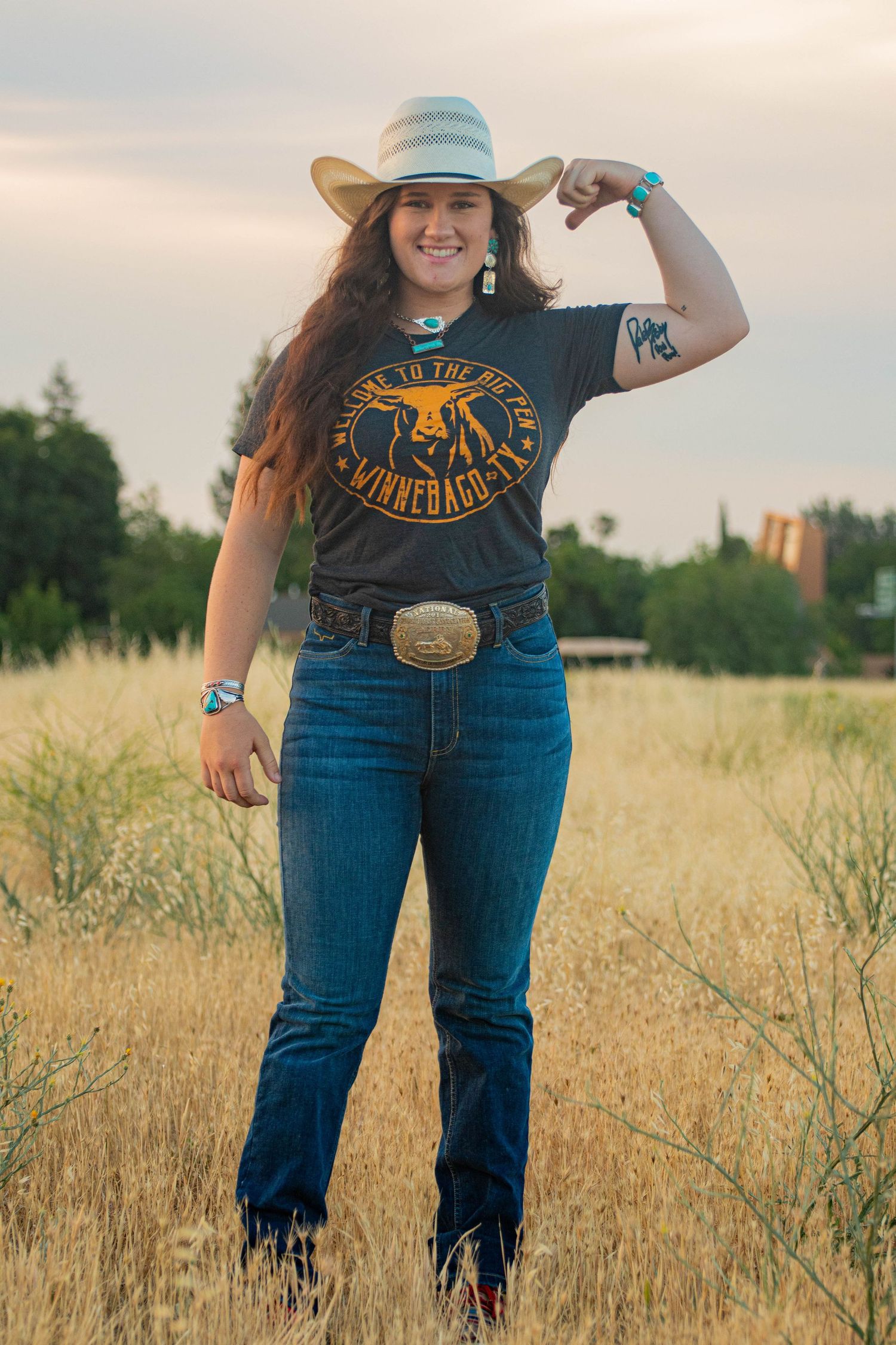 Smiling woman in cowboy hat and jeans flexes arm in golden field wearing Winnebago t-shirt.