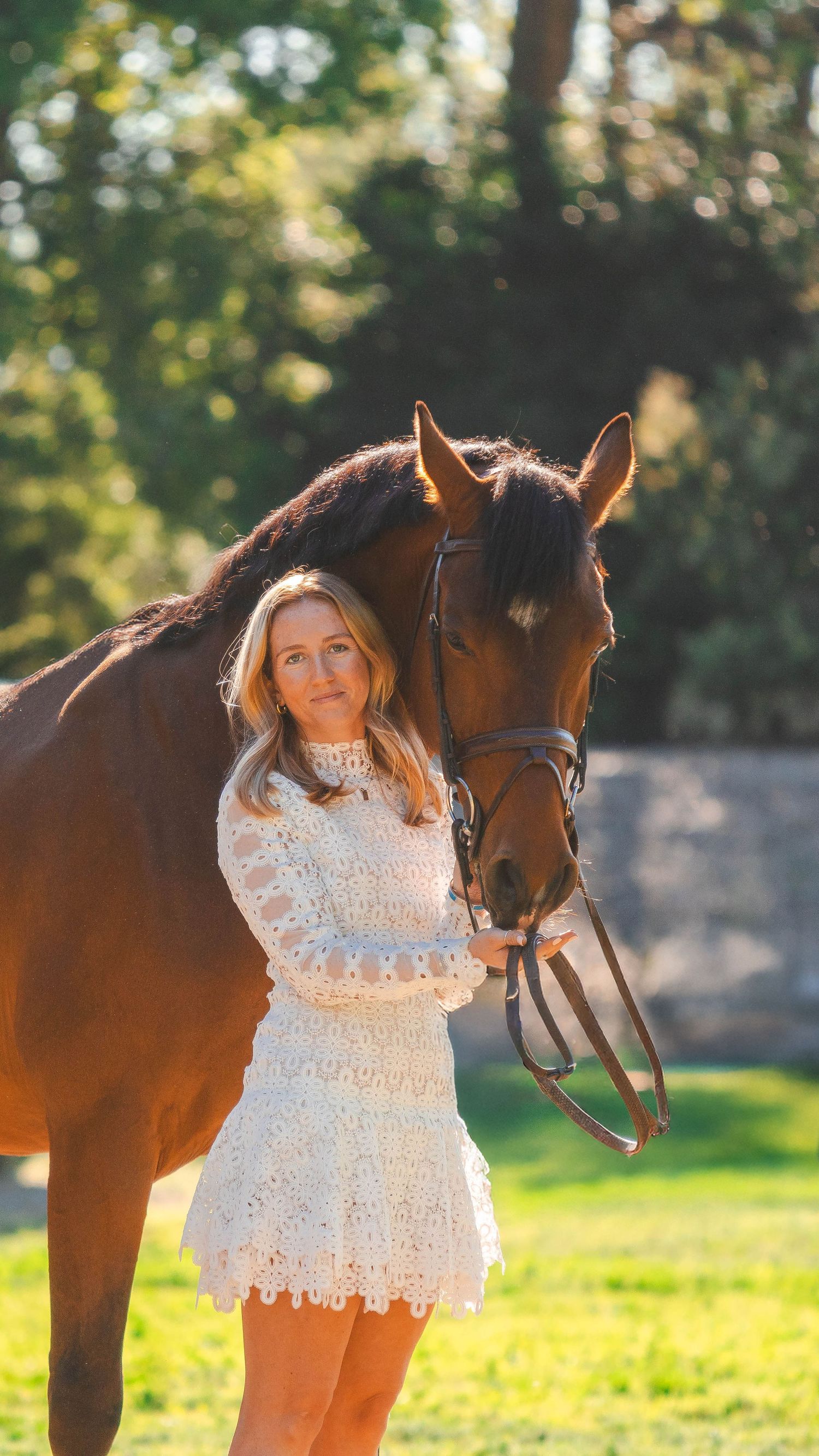 Young blonde woman in white lace dress poses with brown horse in sunny outdoor setting.