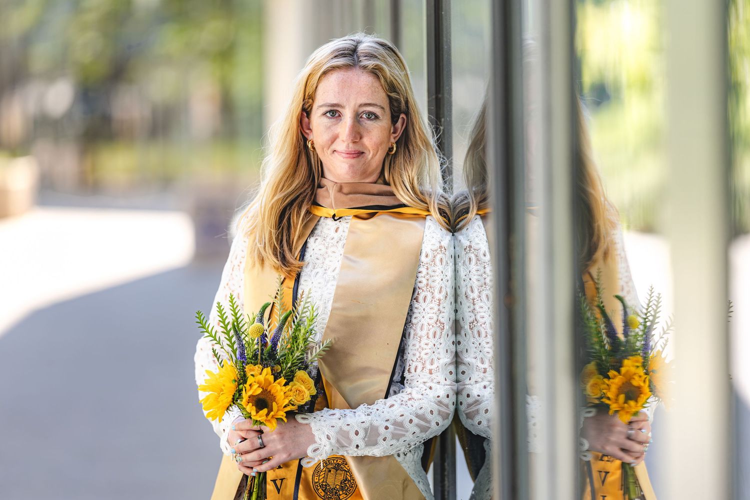 Woman in yellow dress and white lace cardigan holding bright yellow sunflowers, standing outdoors near glass doors.