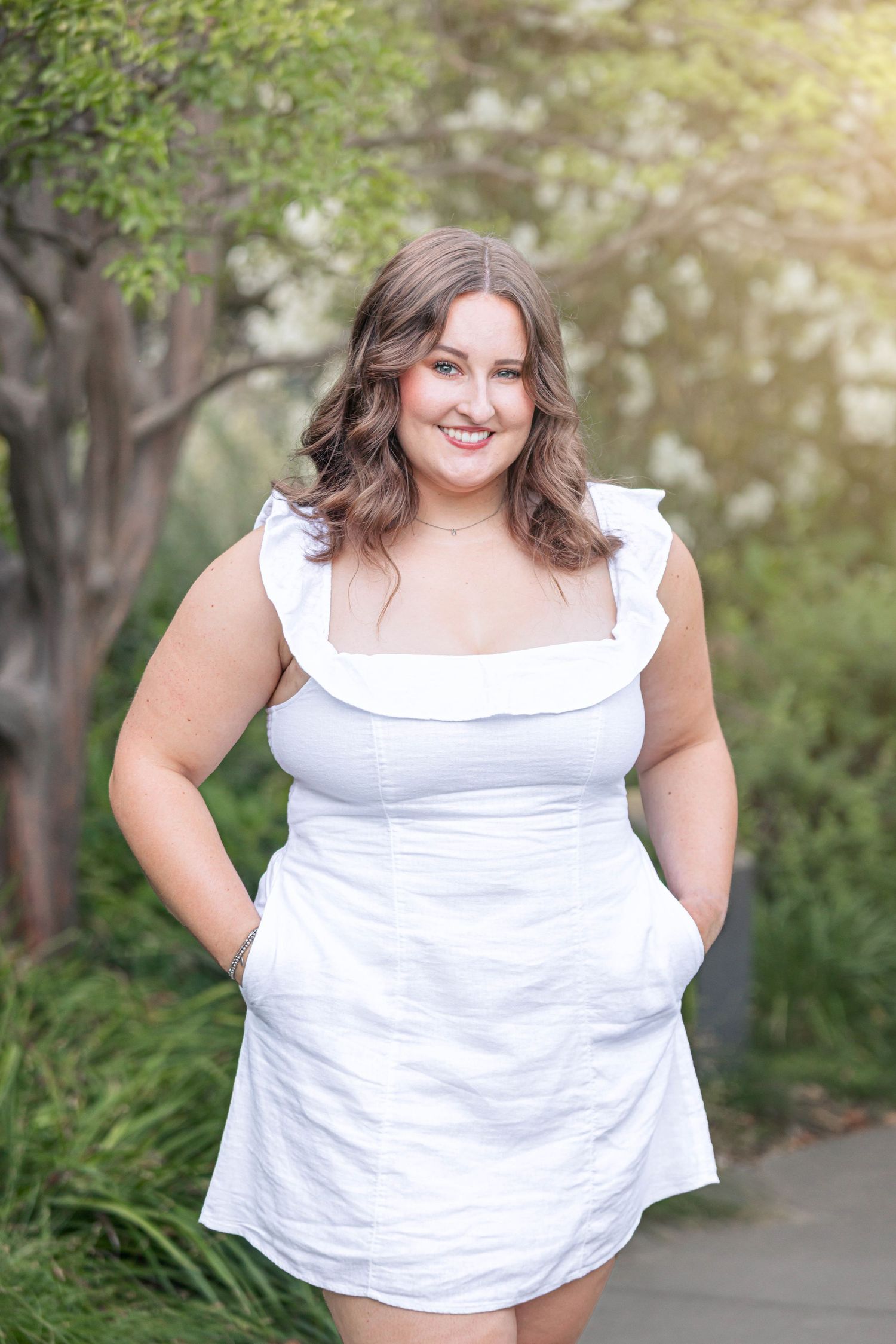 A smiling young woman in a white ruffled mini dress poses outdoors with hands in pockets, surrounded by lush greenery.