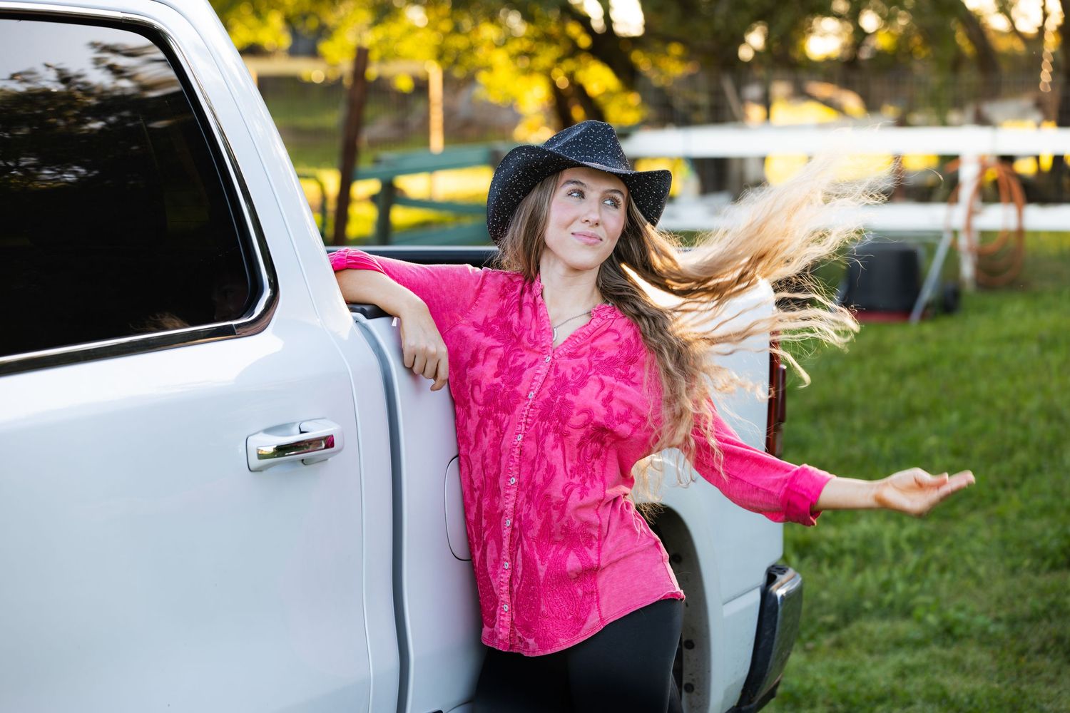 Smiling woman in pink shirt and black cowboy hat posing beside white pickup truck outdoors.