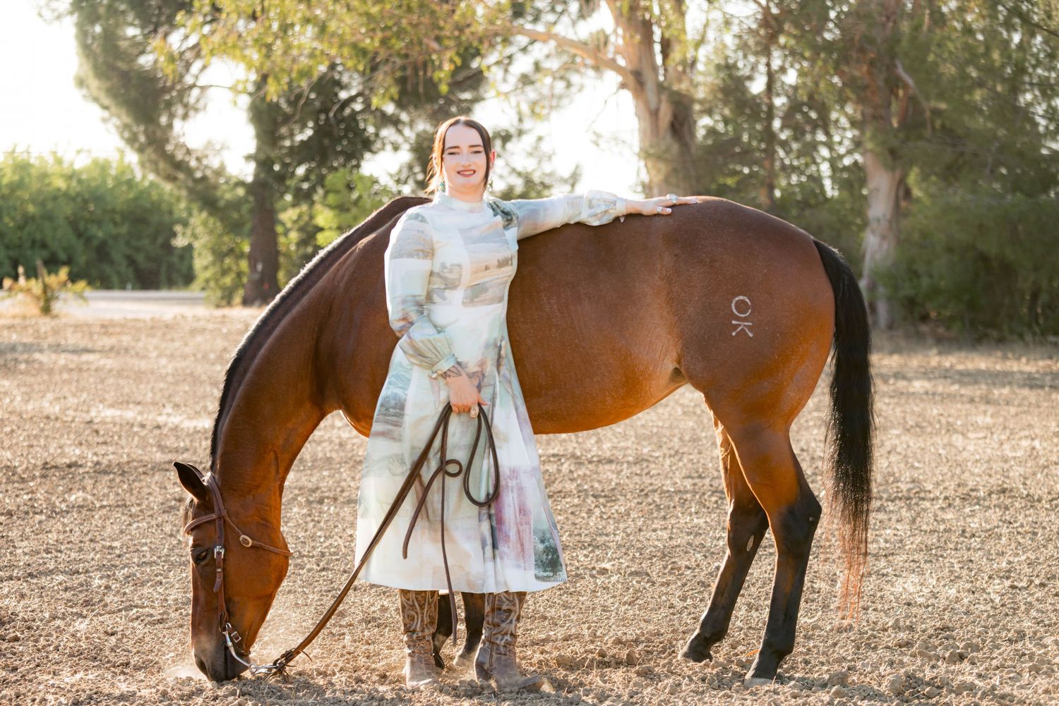 Young woman in pastel dress standing beside a grazing chestnut horse in a sunny outdoor setting.