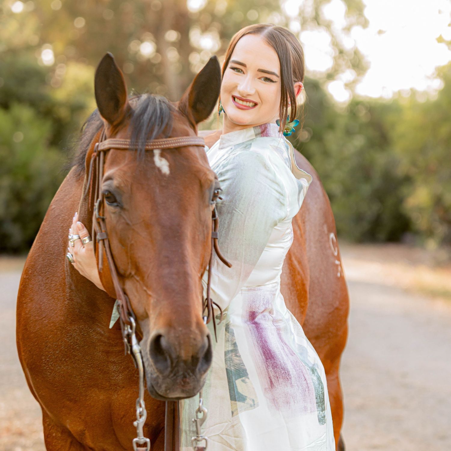 Smiling woman with teal earrings posing beside a chestnut horse on a sunny trail.