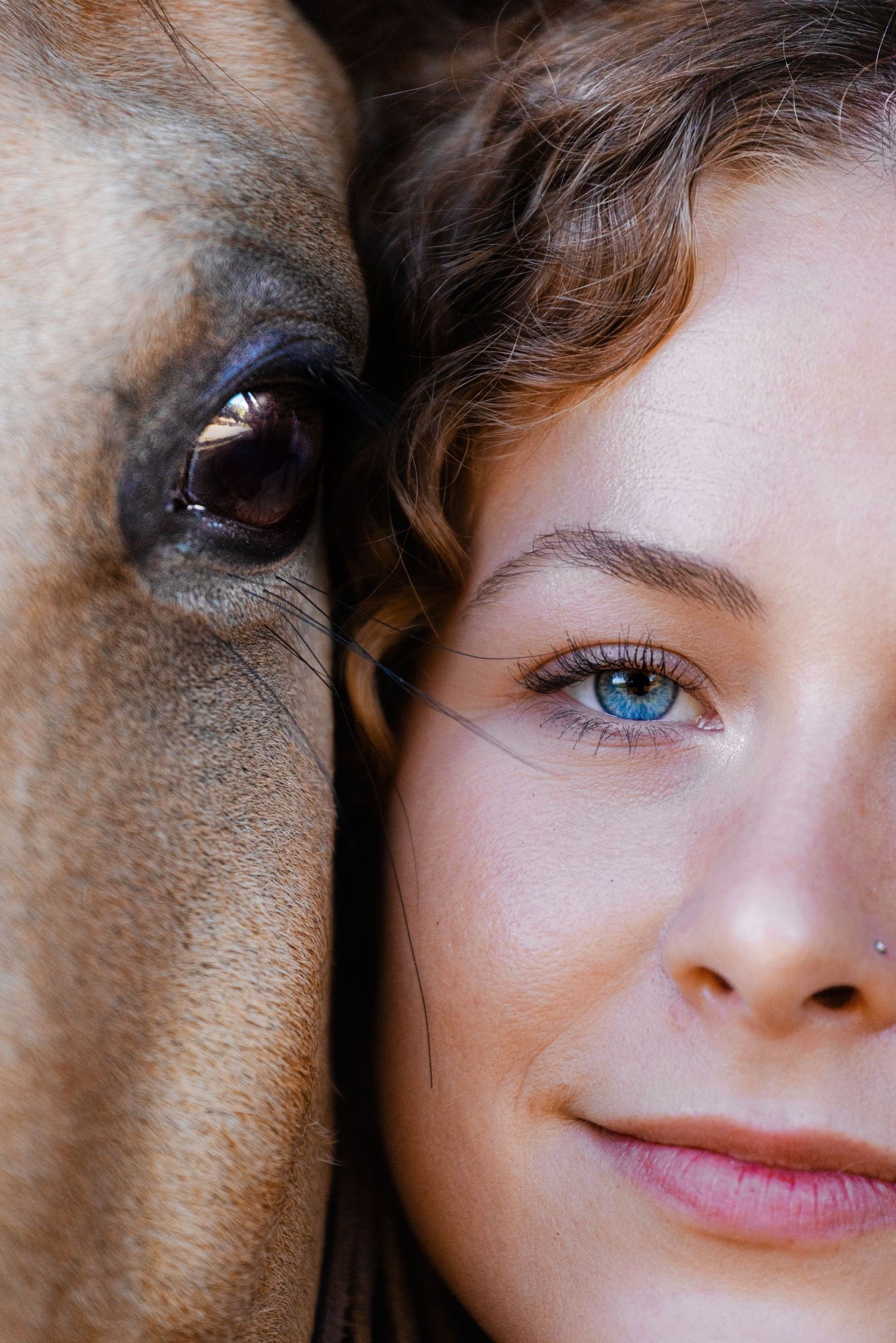 Close-up portrait of blue-eyed woman beside a horse, their faces side by side in intimate moment.