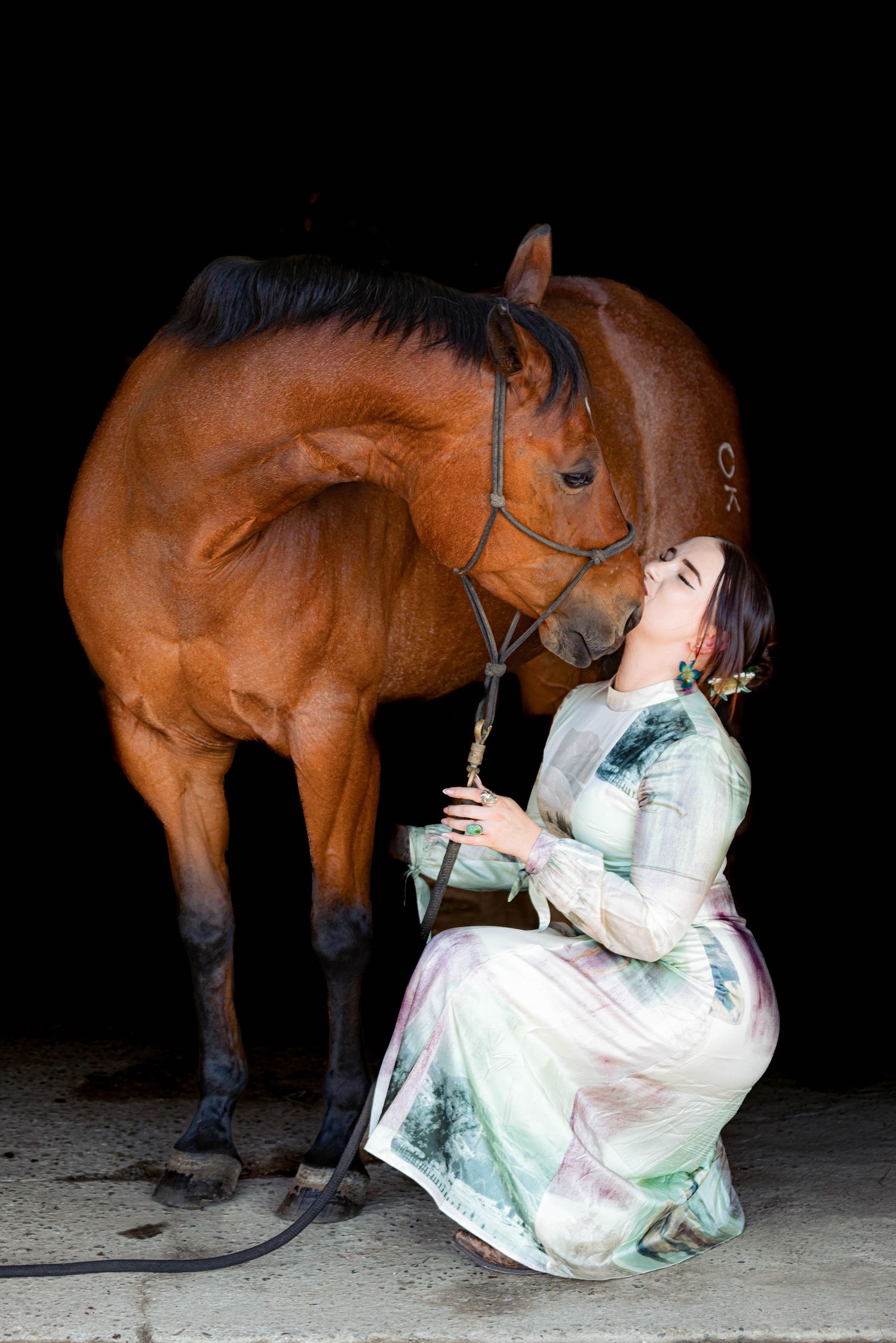 Woman in elegant floral dress sitting beside a bay horse against a dark background.