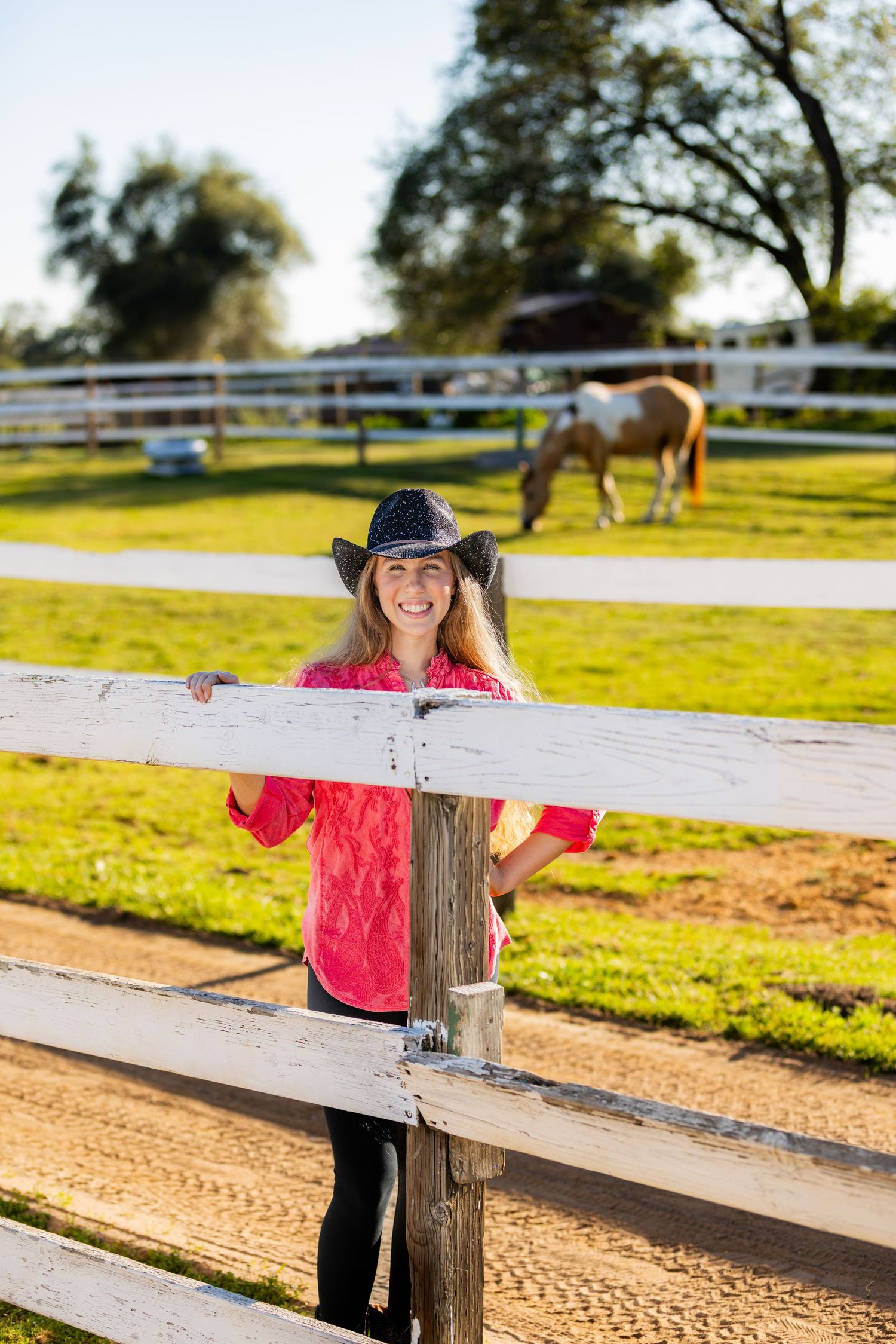 Smiling woman in cowboy hat and pink shirt leaning on white ranch fence with horses grazing in background.
