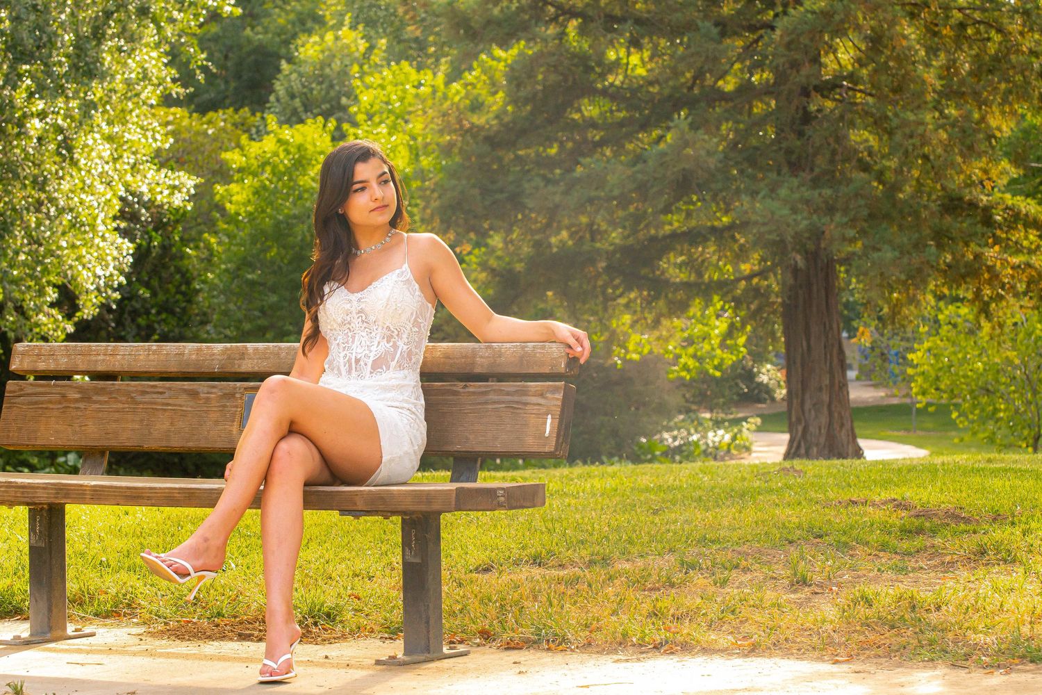 Young woman in white dress sitting on a park bench surrounded by sunny green trees.