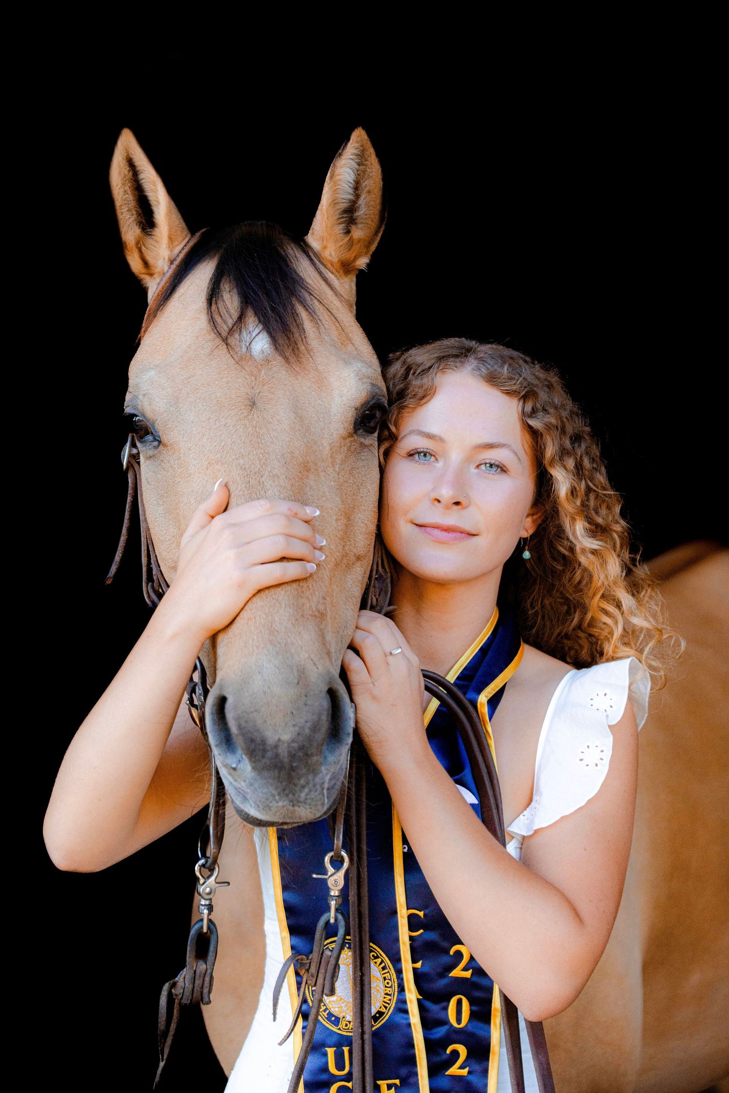 Young woman with curly hair wearing a blue sash poses with a buckskin horse against a black background.