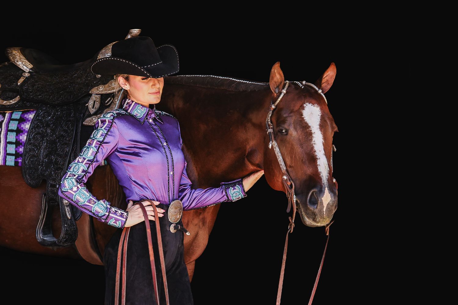 Woman in purple western show shirt and black cowboy hat poses with chestnut horse against dark background.