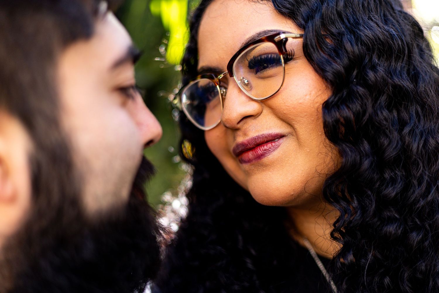 Close-up of couple gazing at each other, woman wearing glasses smiling warmly outdoors.