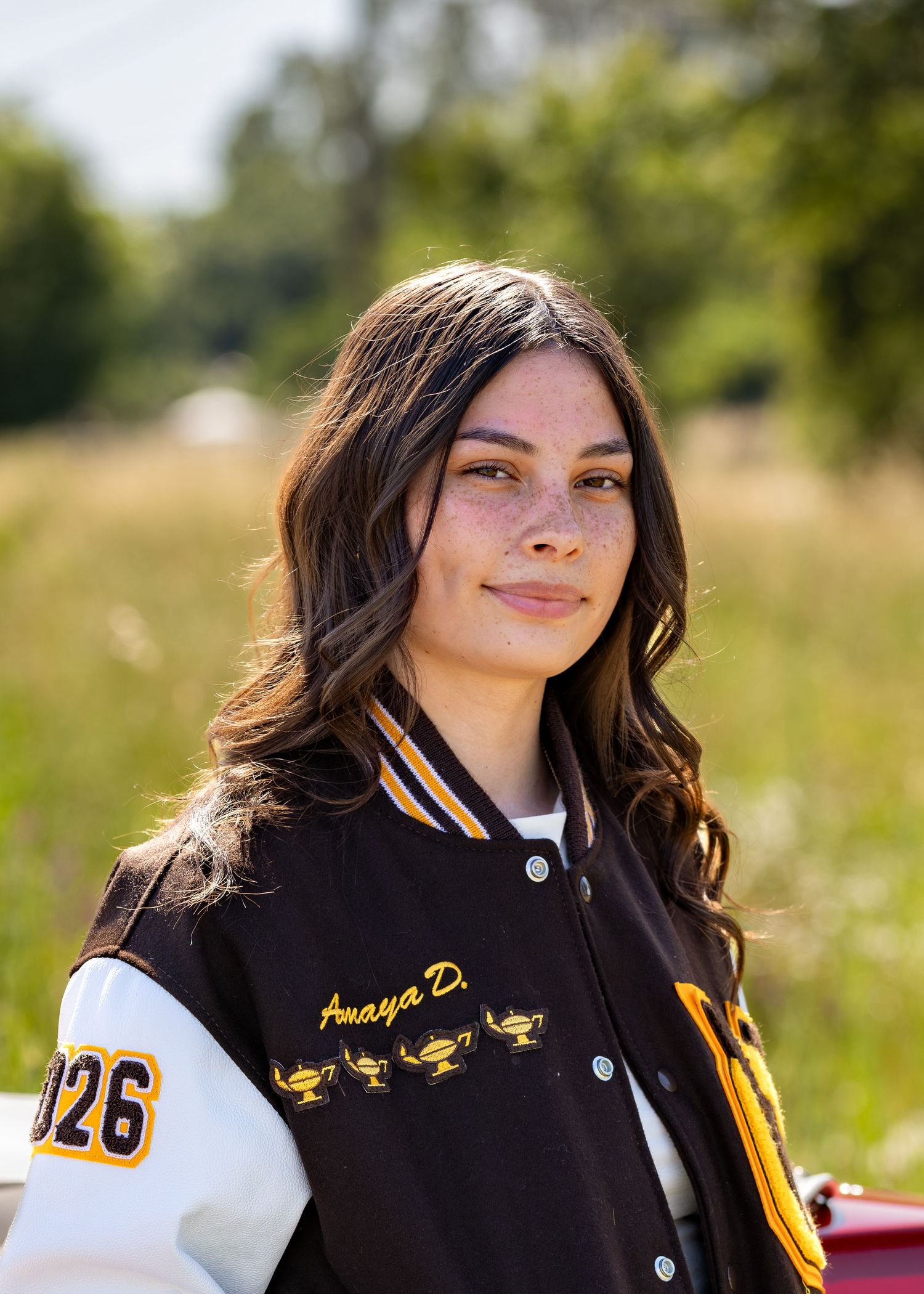 Young woman in black varsity jacket with name Amaya D. smiling outdoors in a sunny field.