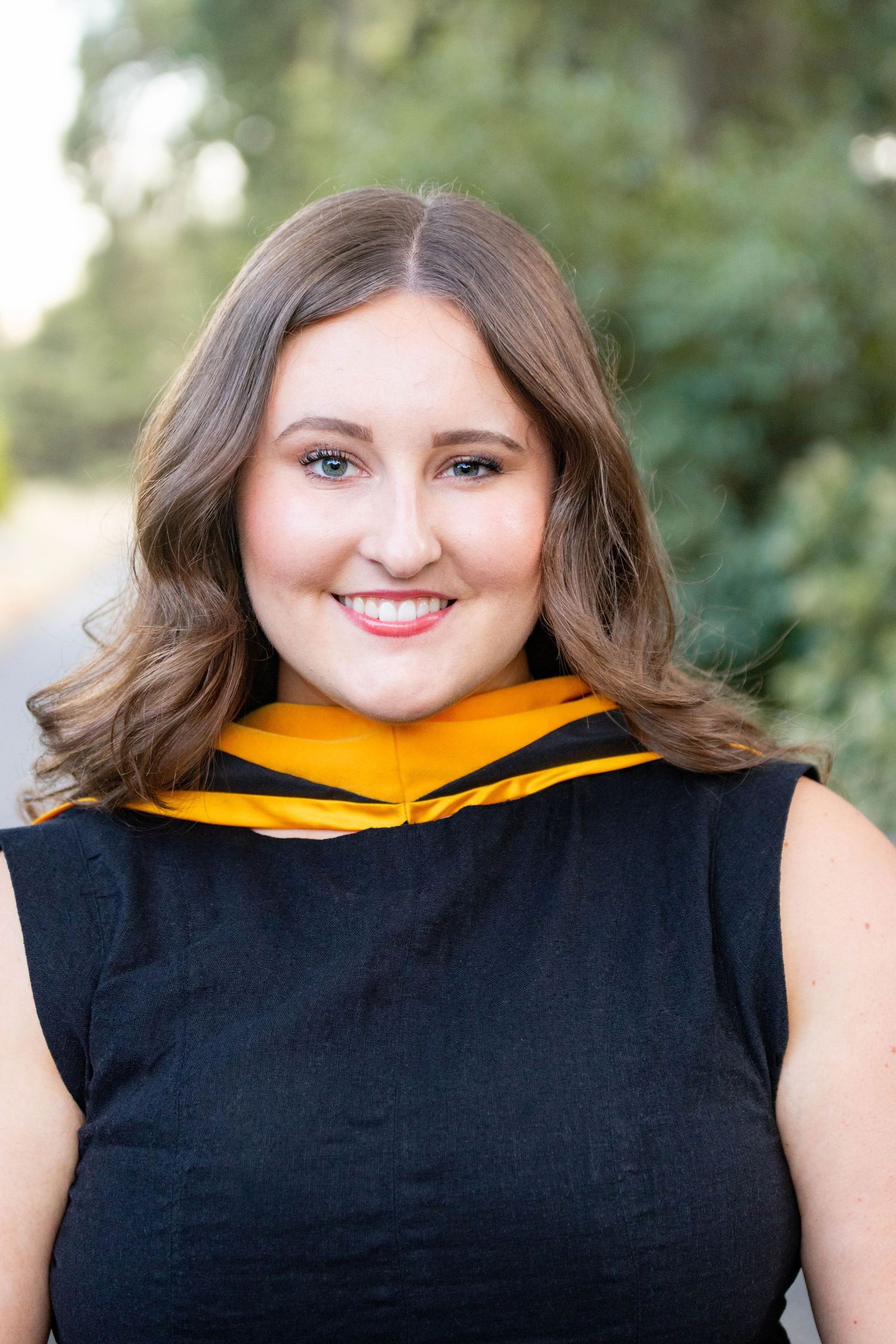 Young woman in black graduation gown with gold hood smiling outdoors with green trees in background.