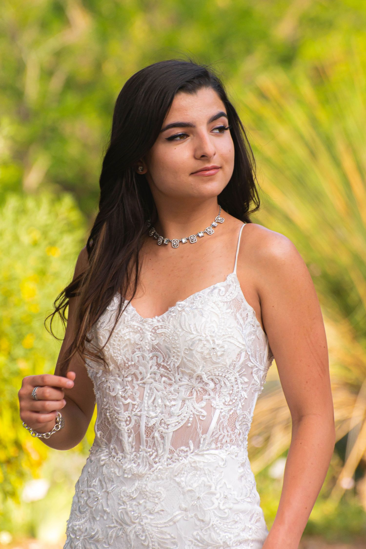 Young woman in white lace dress and diamond necklace posing outdoors with lush green foliage background.