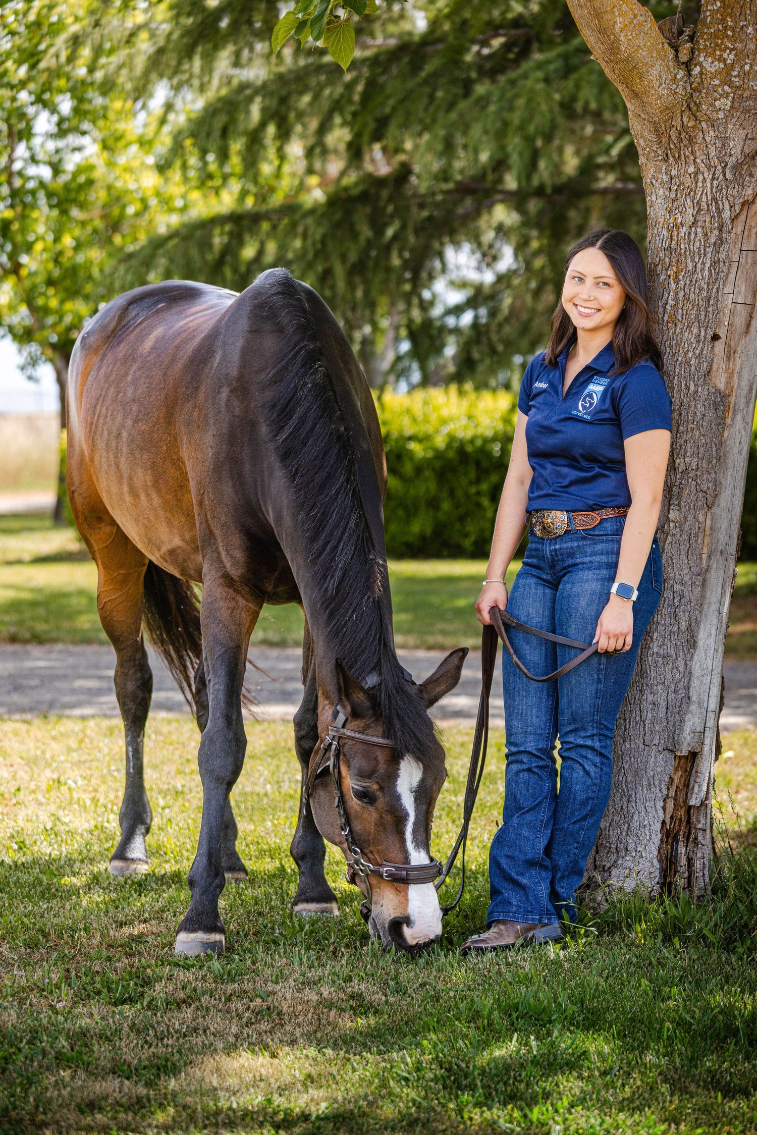 Smiling woman in blue polo shirt standing with a bay horse grazing under a tree outdoors.