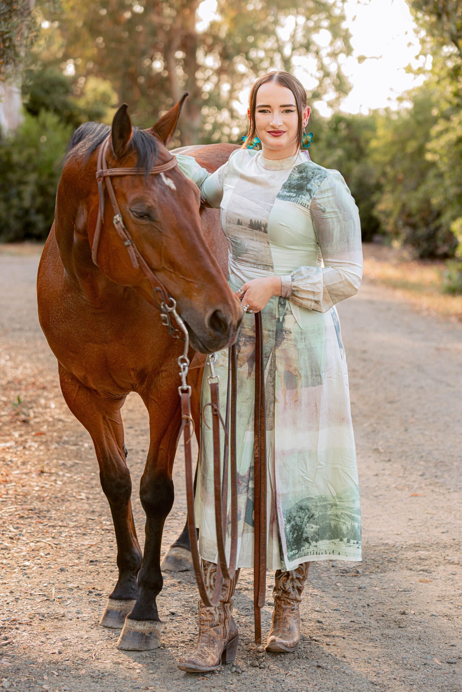 Woman in sheer floral dress standing with a bay horse on a dirt path outdoors.