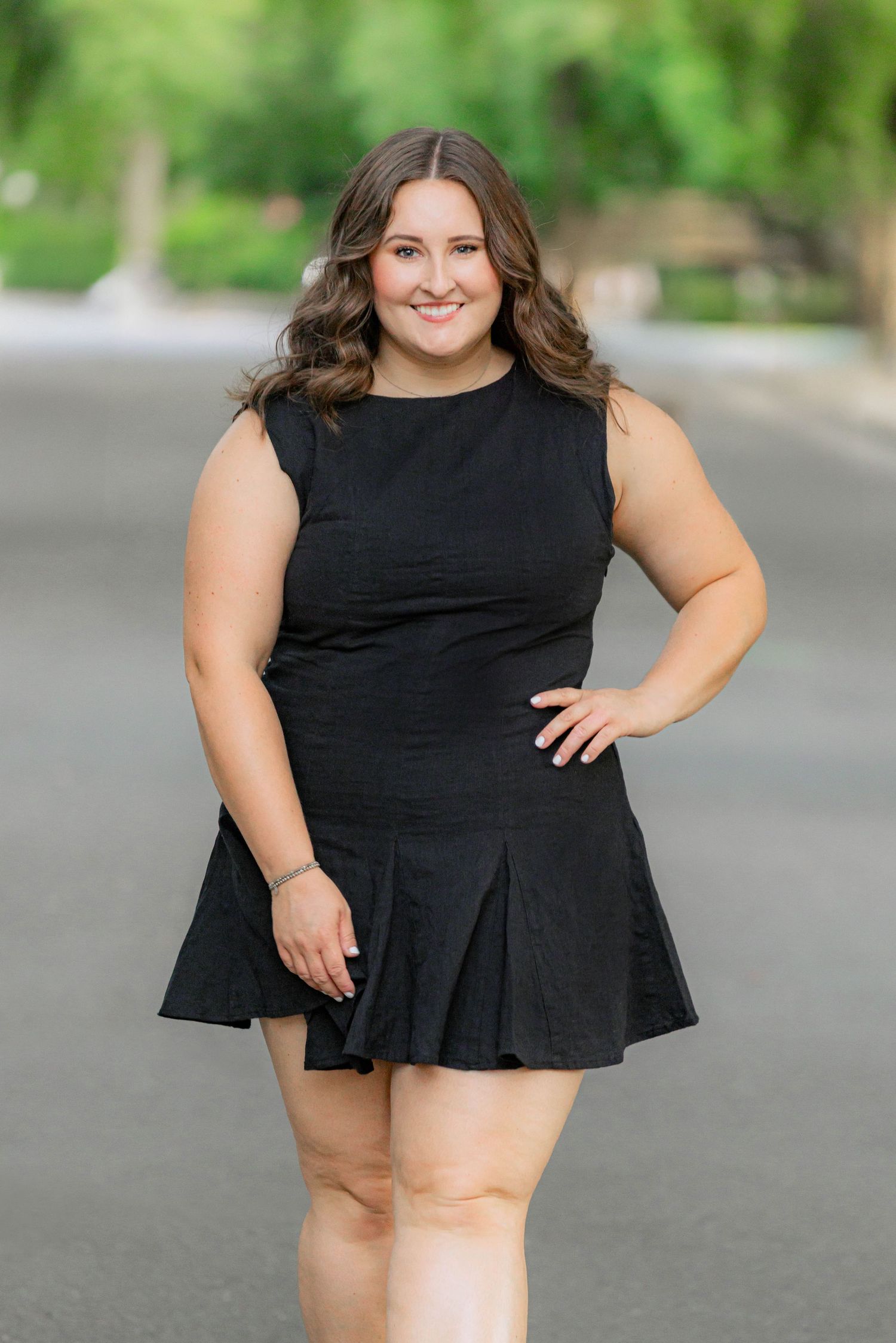 A smiling young woman in a black sleeveless mini dress poses confidently on a tree-lined street.