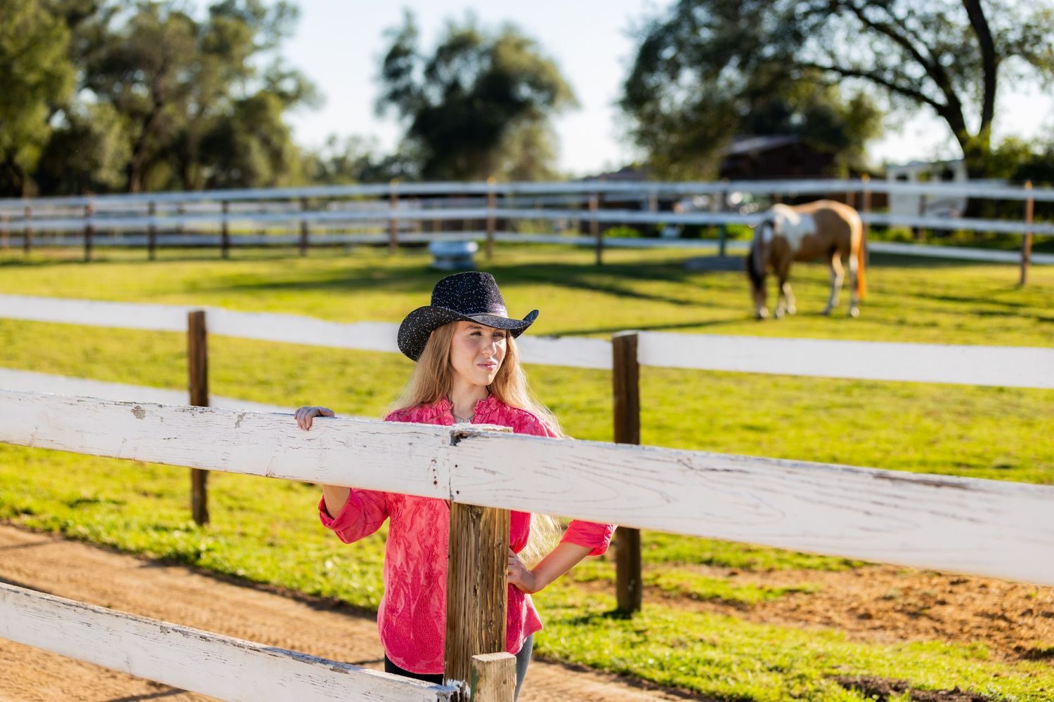 Young blonde woman in pink shirt and cowboy hat leaning on white ranch fence with horse grazing in background.
