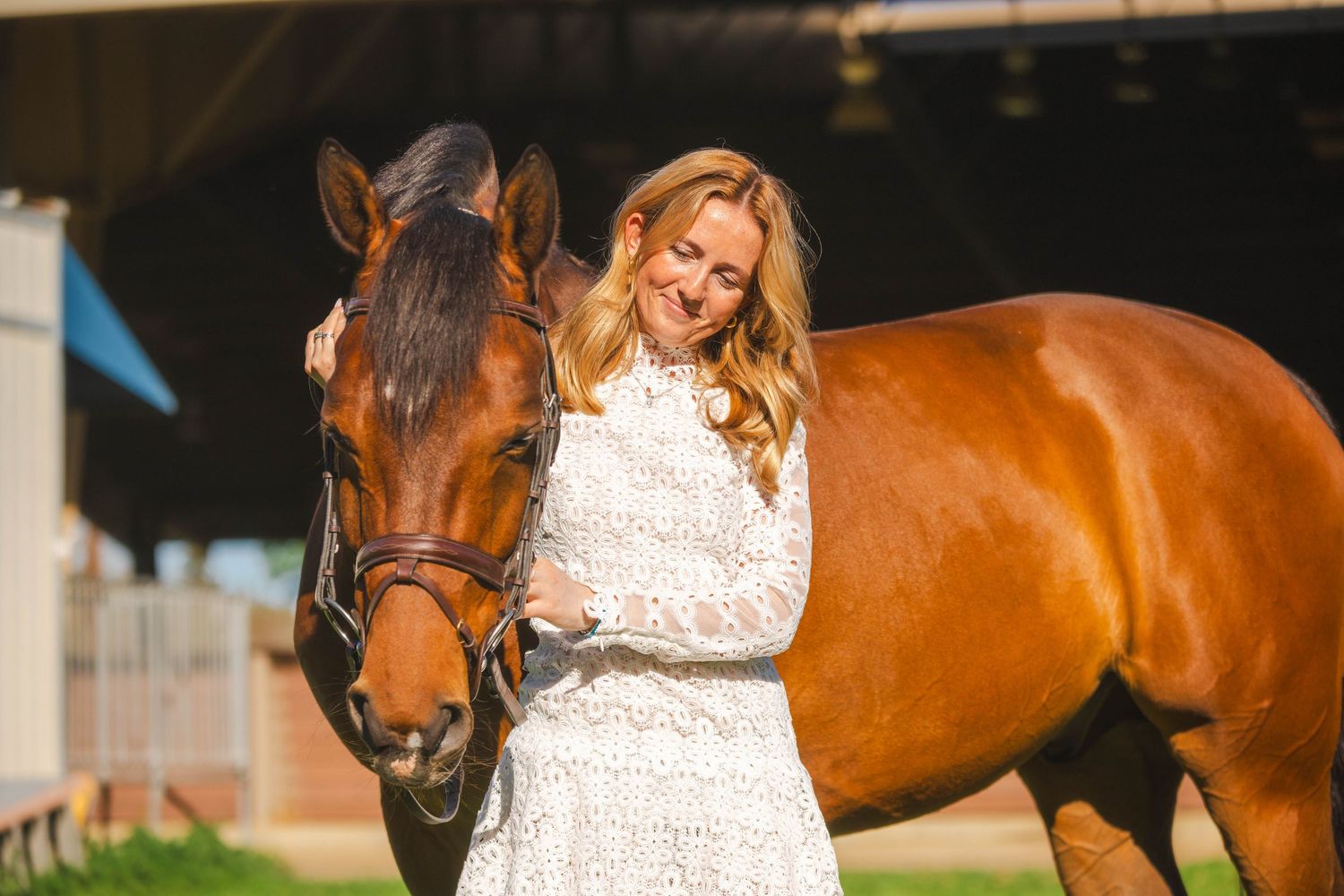 Smiling blonde woman in white lace dress stands beside chestnut horse near stable barn.