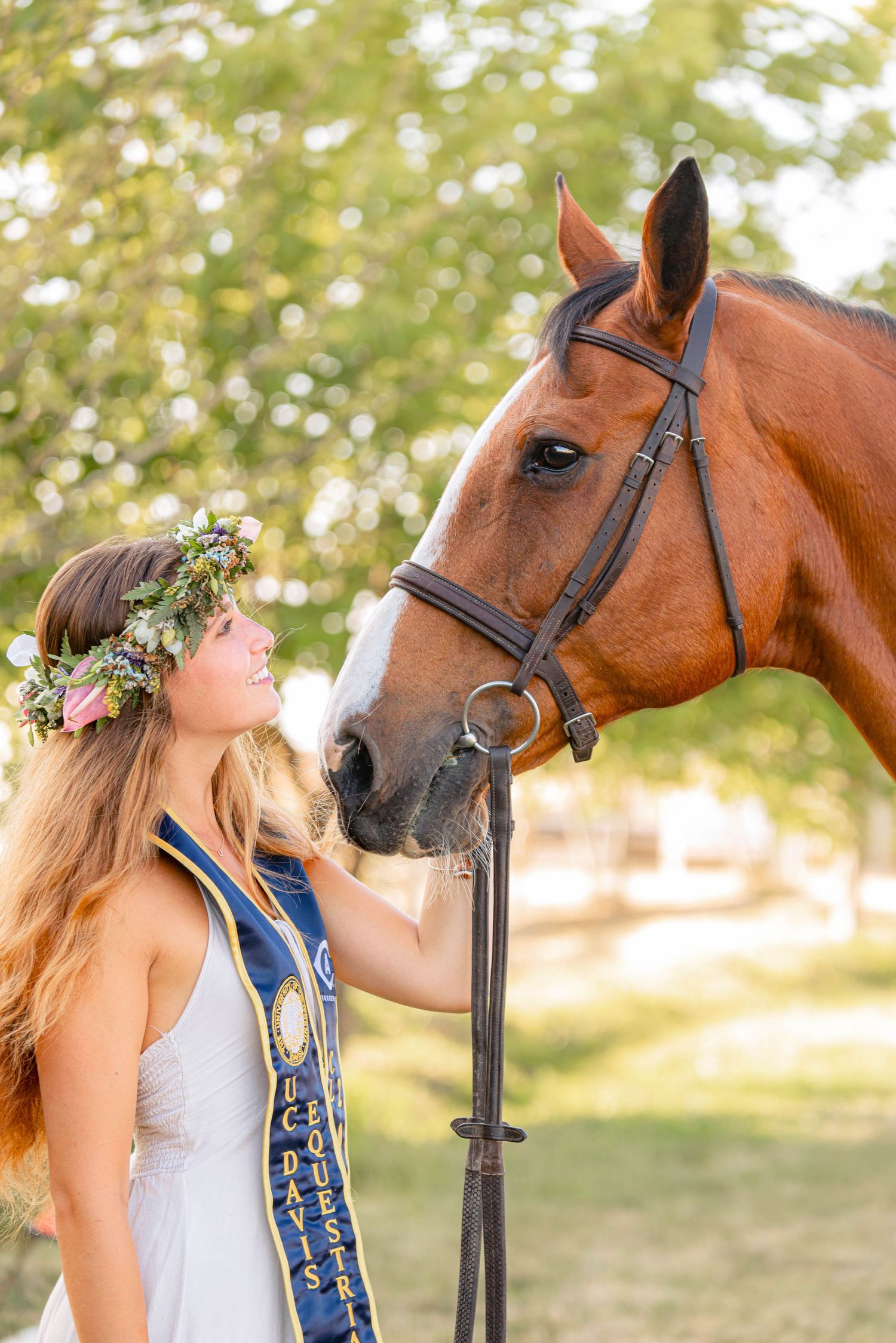 Young woman with flower crown and equestrian sash poses with a bay horse outdoors.