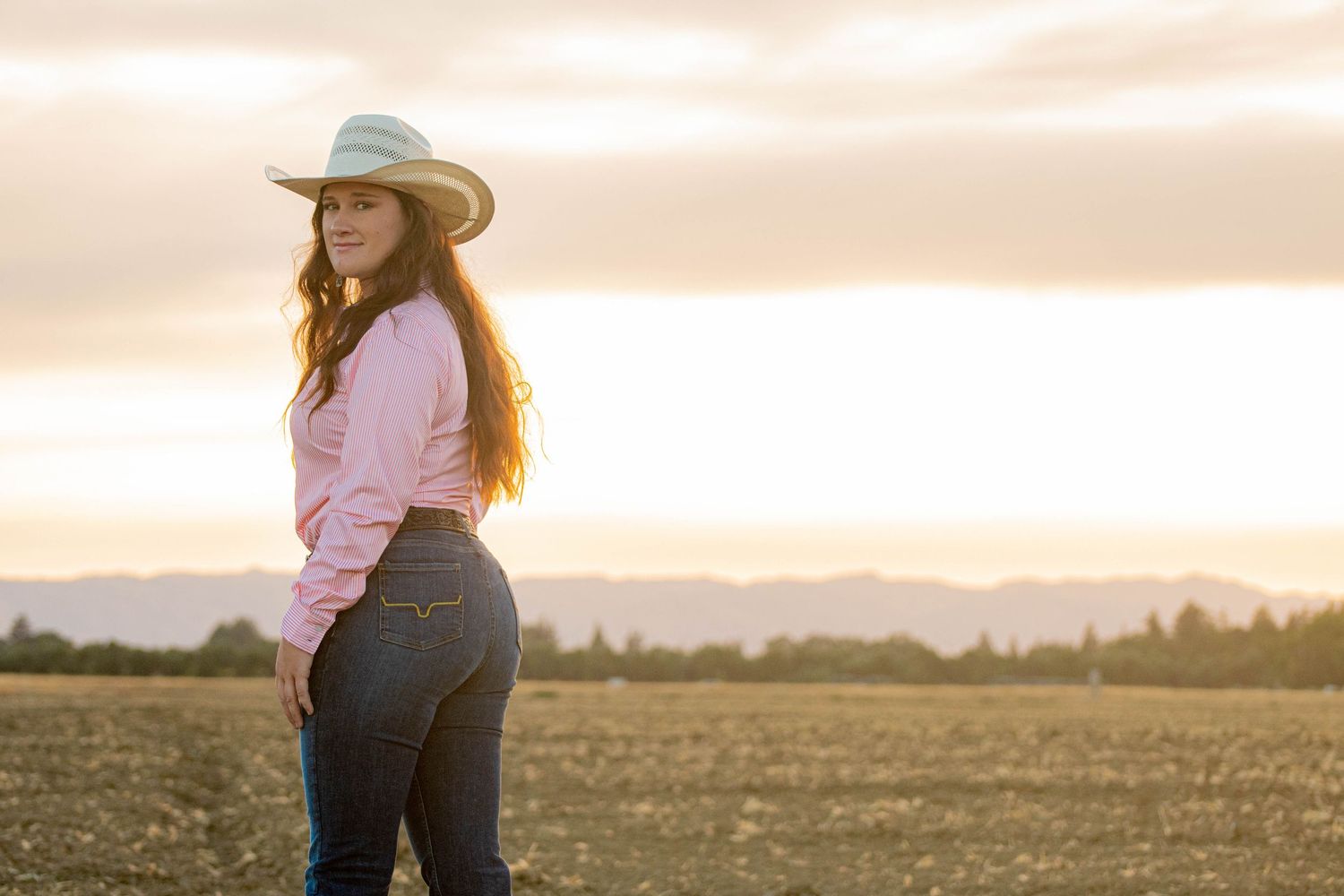 Young woman in pink shirt, jeans, and cowboy hat poses in a field at golden sunset.