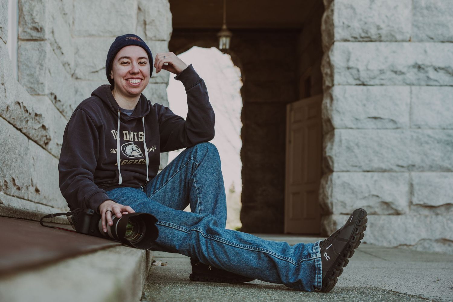 Smiling young person in hoodie and jeans sitting on steps, flexing arm muscles outdoors near stone building.