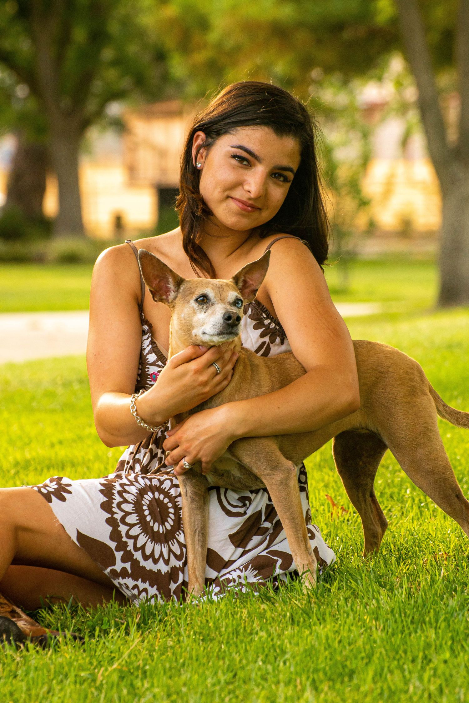 Young woman in floral dress sitting on green grass in a park, holding a small tan Chihuahua mix dog.