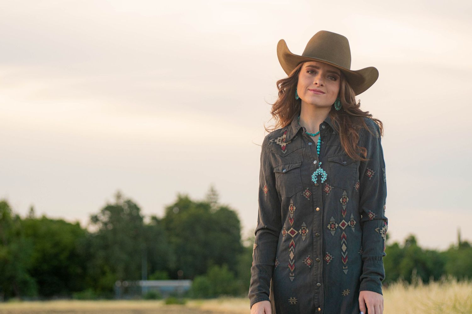 Woman in cowboy hat and southwestern-patterned jacket with turquoise necklace standing in open field.