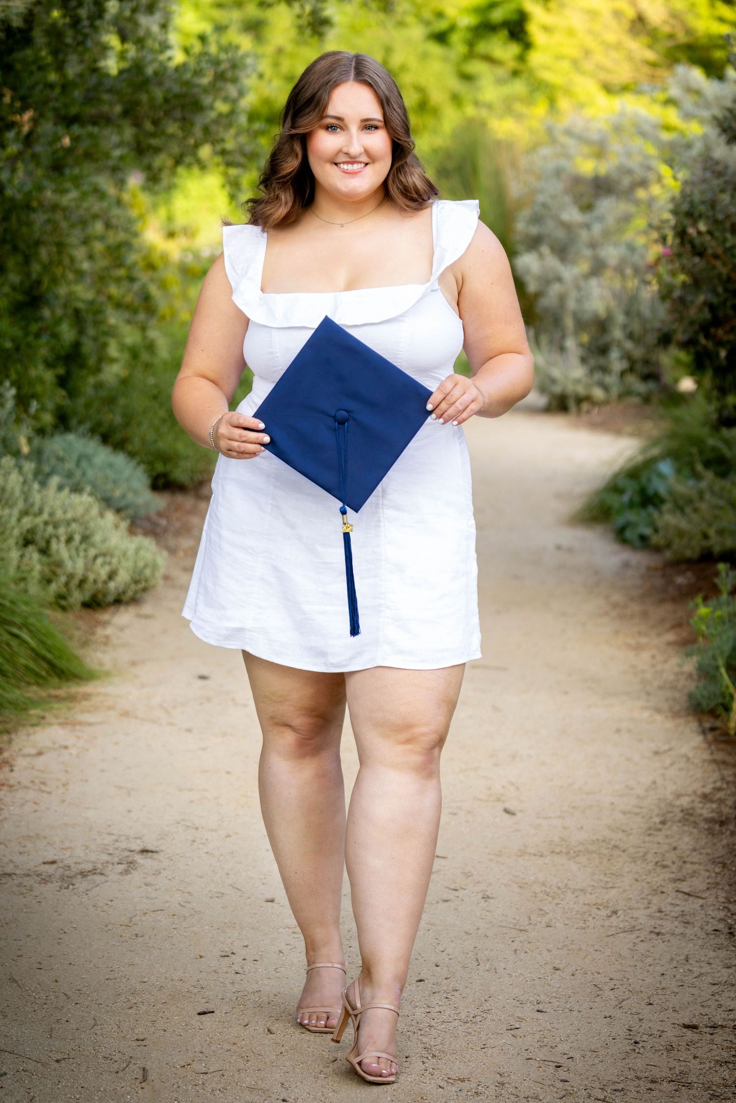 Smiling young woman in white dress holding navy blue graduation cap while walking on a garden path.