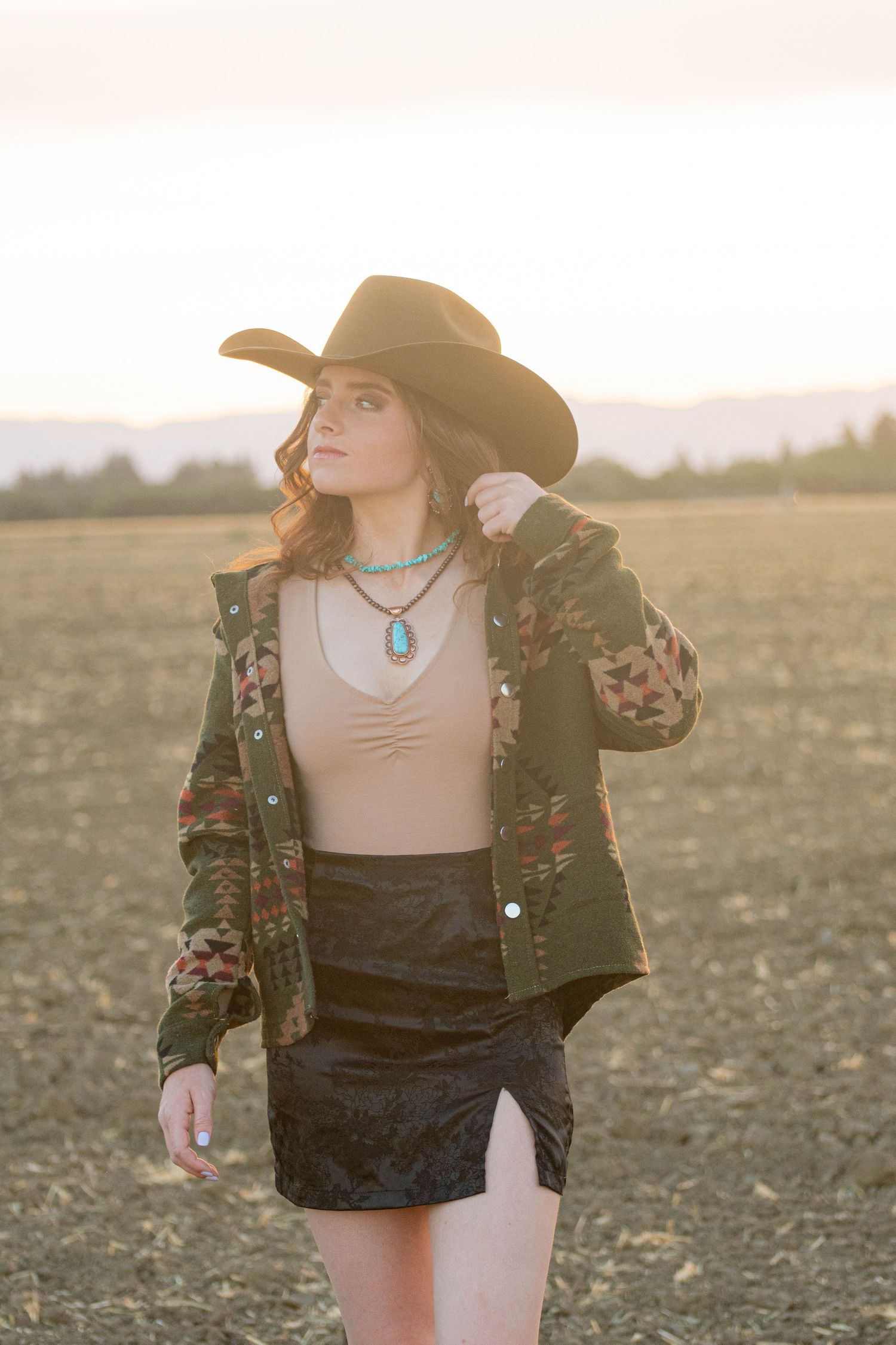 Stylish woman adjusting black cowboy hat wearing camo jacket and turquoise pendant necklace in golden hour outdoor field.