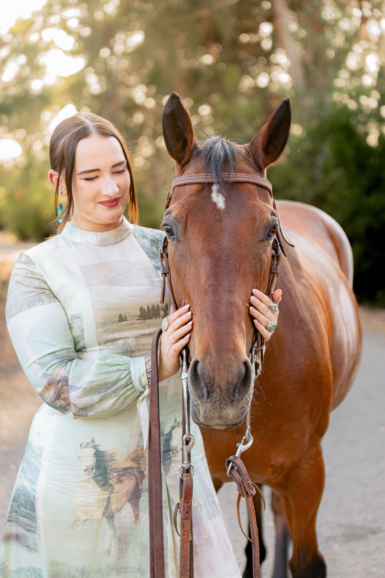Smiling brunette woman in green print dress posing with a chestnut horse outdoors.
