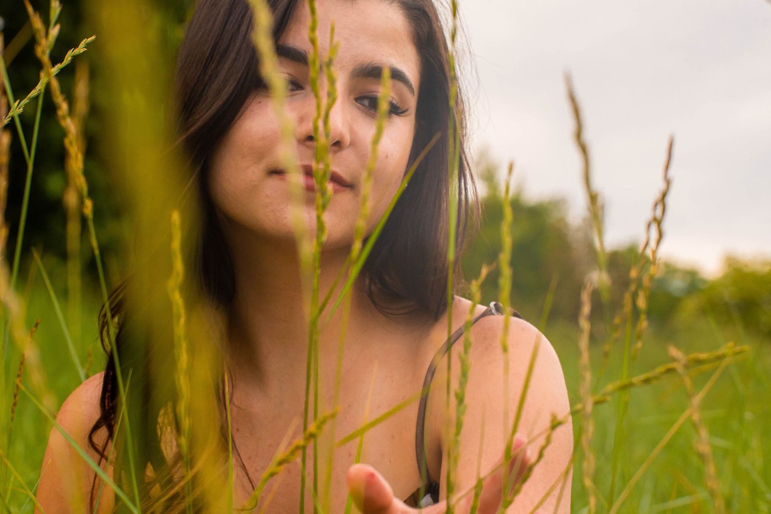 Young woman with dark hair standing in tall green grass field, reaching out her hand in a serene outdoor setting.