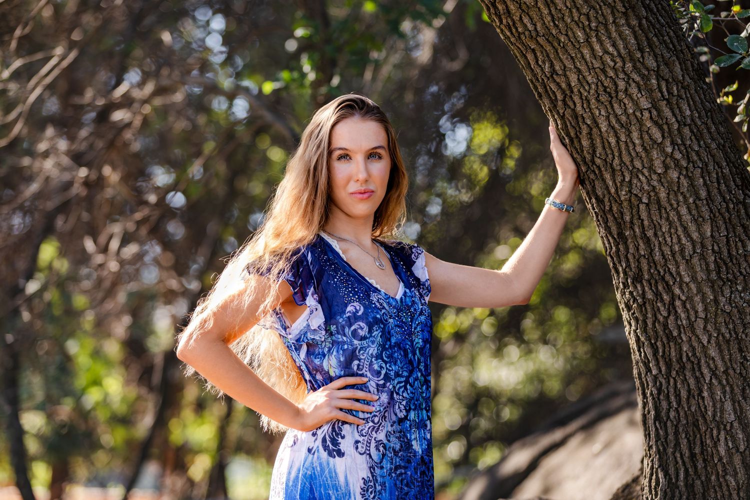 A woman in a blue floral dress poses gracefully beside a tree in a sunlit outdoor park setting.