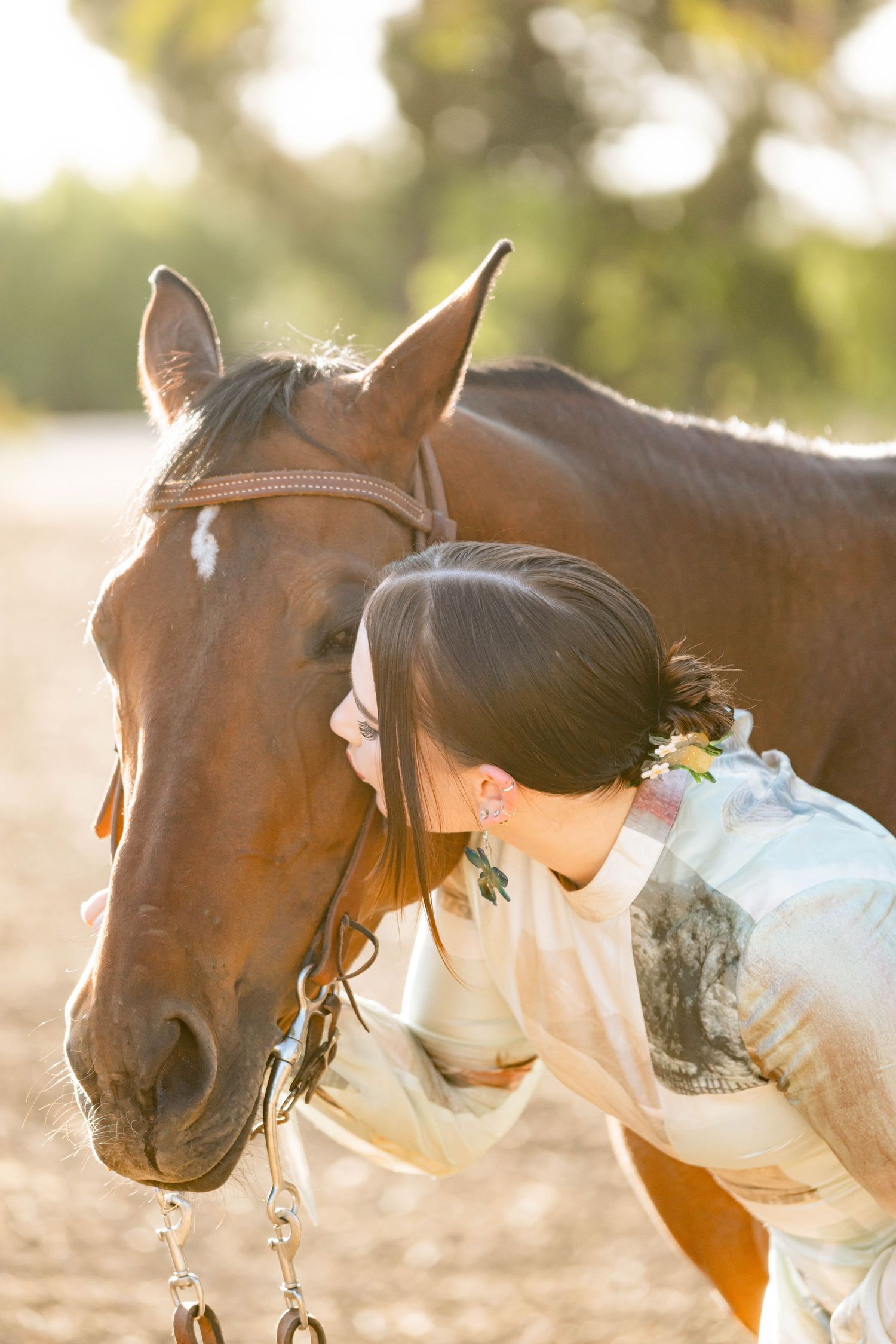 Young woman tenderly kissing a chestnut horse on the nose in warm golden sunlight outdoors.