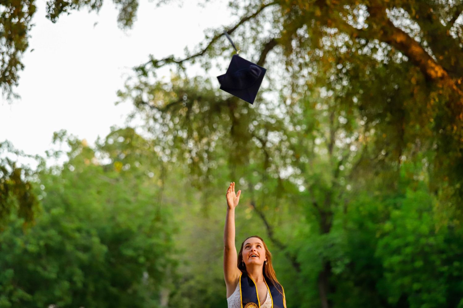 Joyful graduate tossing navy graduation cap into air surrounded by lush green trees outdoors.