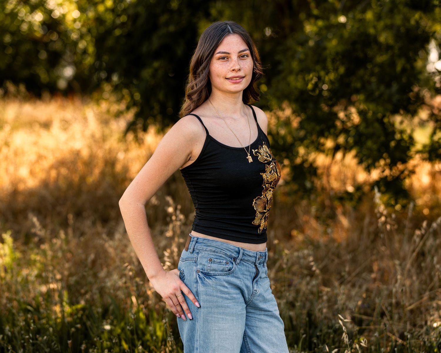 Young woman in black crop top and jeans poses outdoors in golden sunlit field with trees.