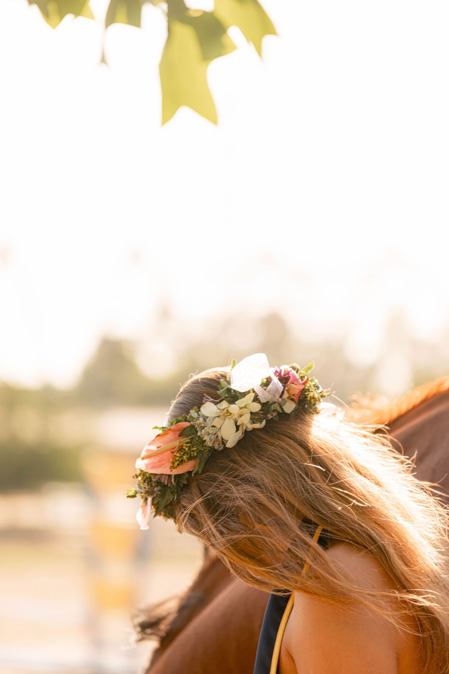 Woman with flowing red hair wearing a floral crown of pink and white flowers, photographed outdoors in warm golden light.