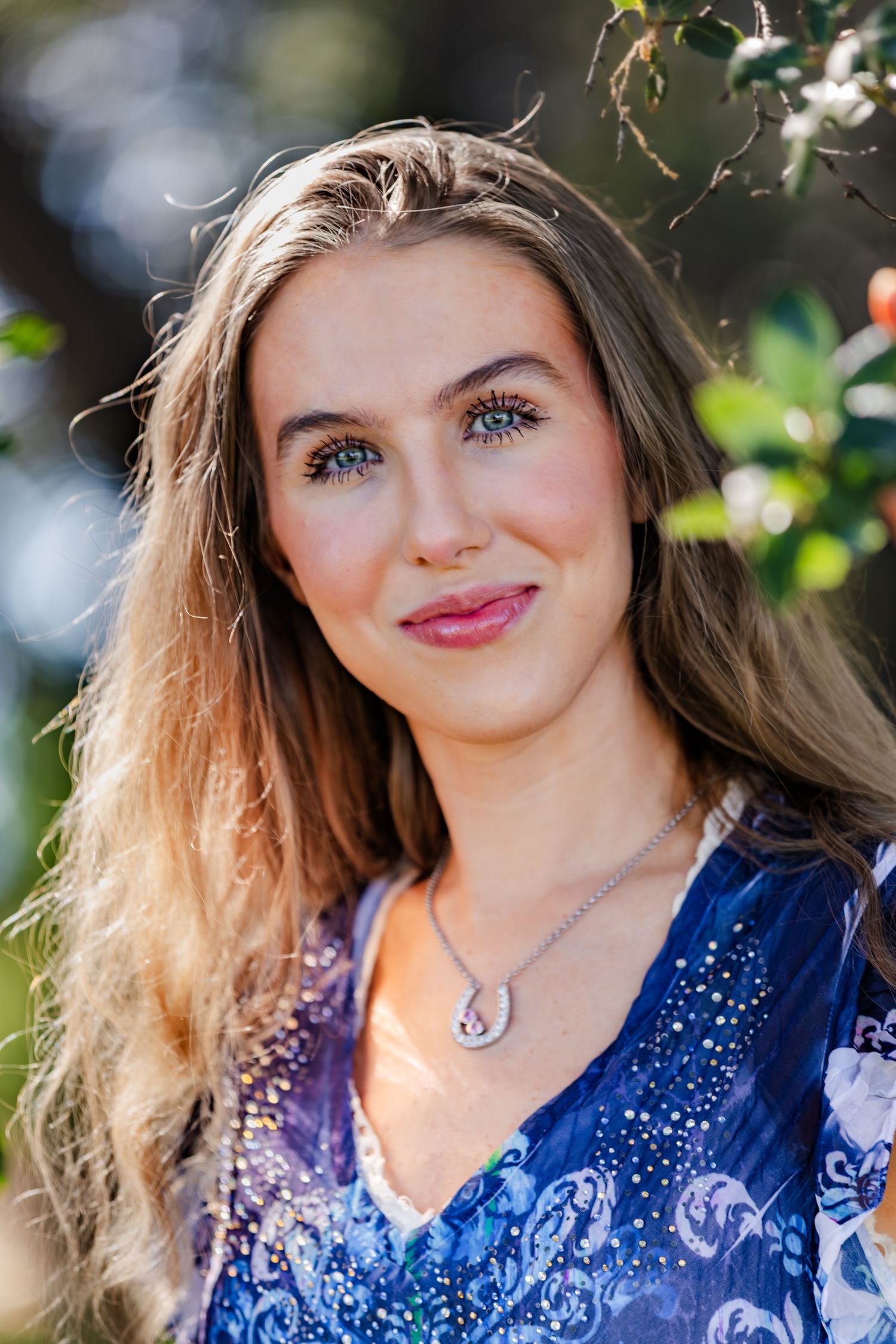 Young woman with long blonde hair and blue eyes wearing a blue floral top and silver necklace outdoors.