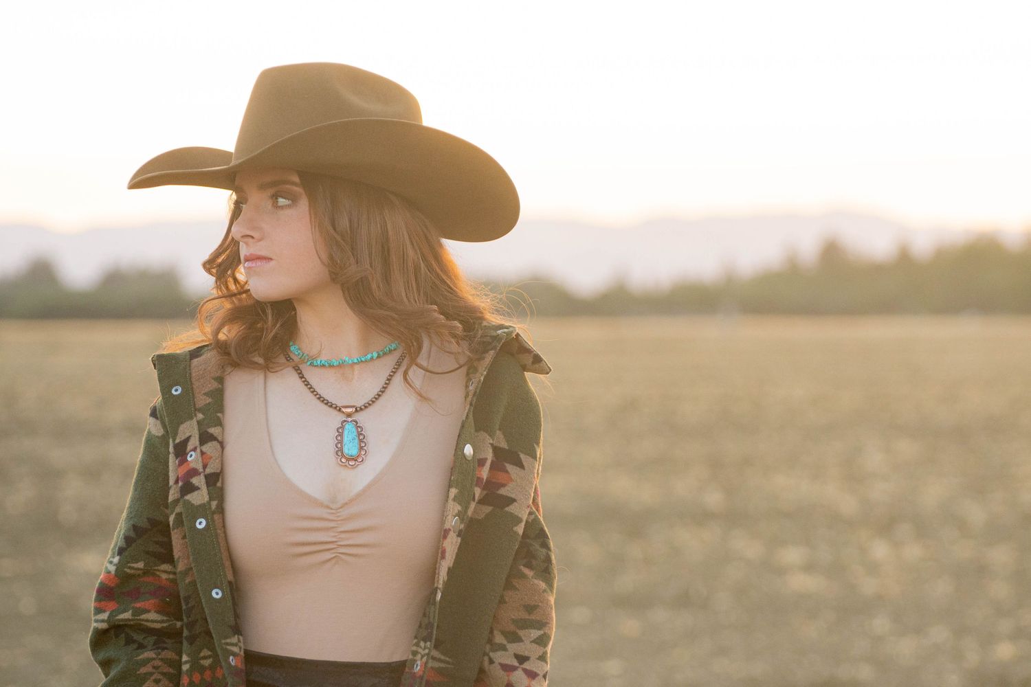 Young woman in black cowboy hat and turquoise necklace standing in golden field at sunset.