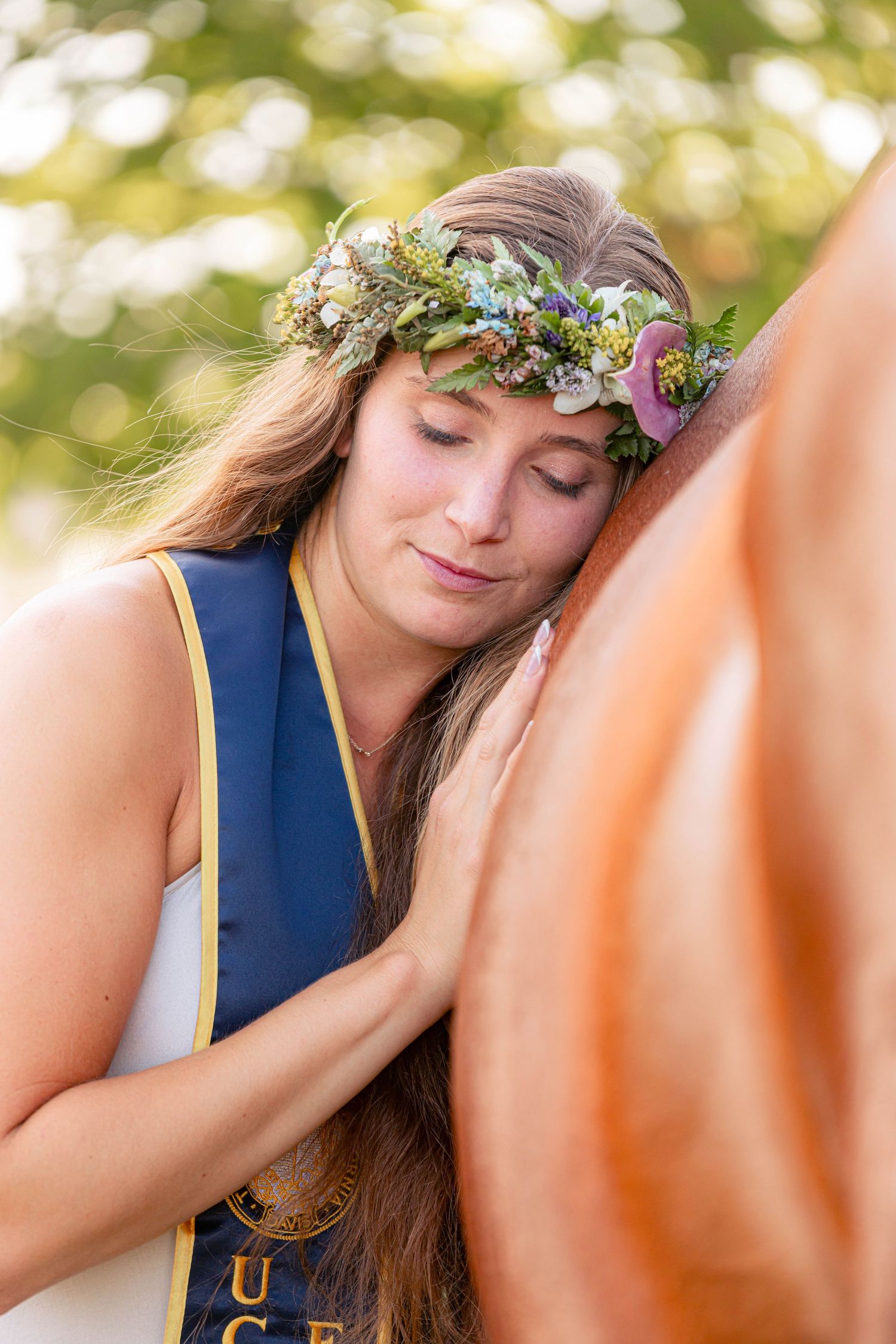 Young woman in graduation attire and floral crown rests her head against a horse outdoors.