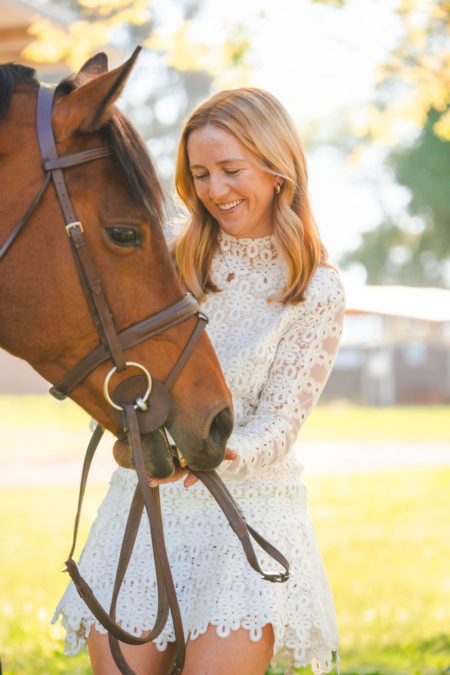 Smiling blonde woman in white lace dress feeding and petting a chestnut horse in sunny field.