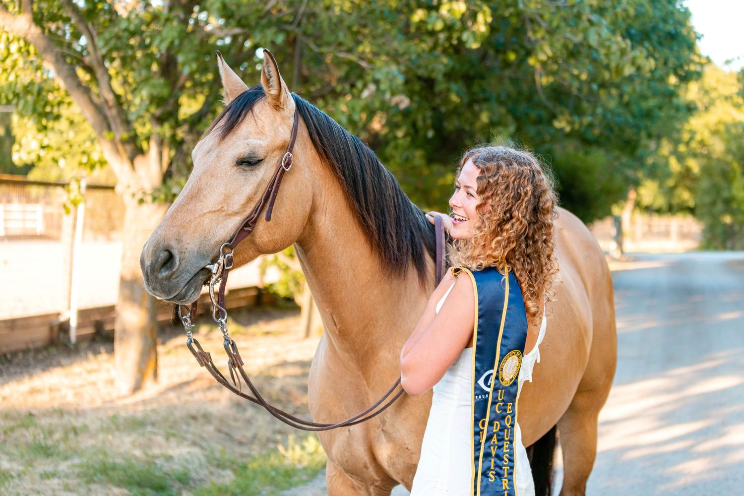 Young woman with curly hair standing beside a buckskin horse displaying a blue ribbon outdoors.