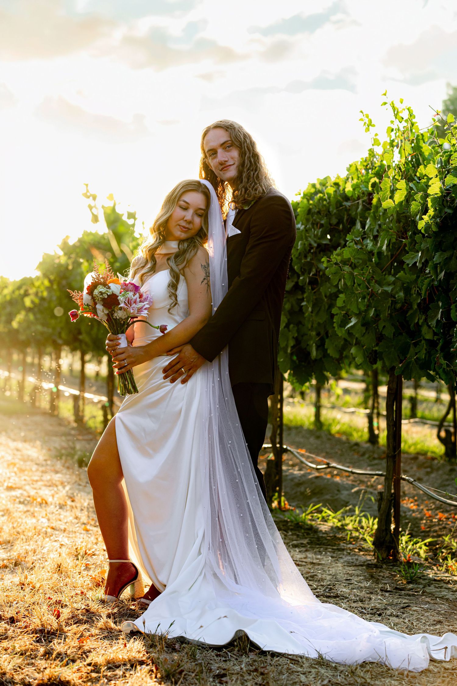Bride and groom embracing in a vineyard at golden hour, bride holding colorful bouquet in white gown with slit.