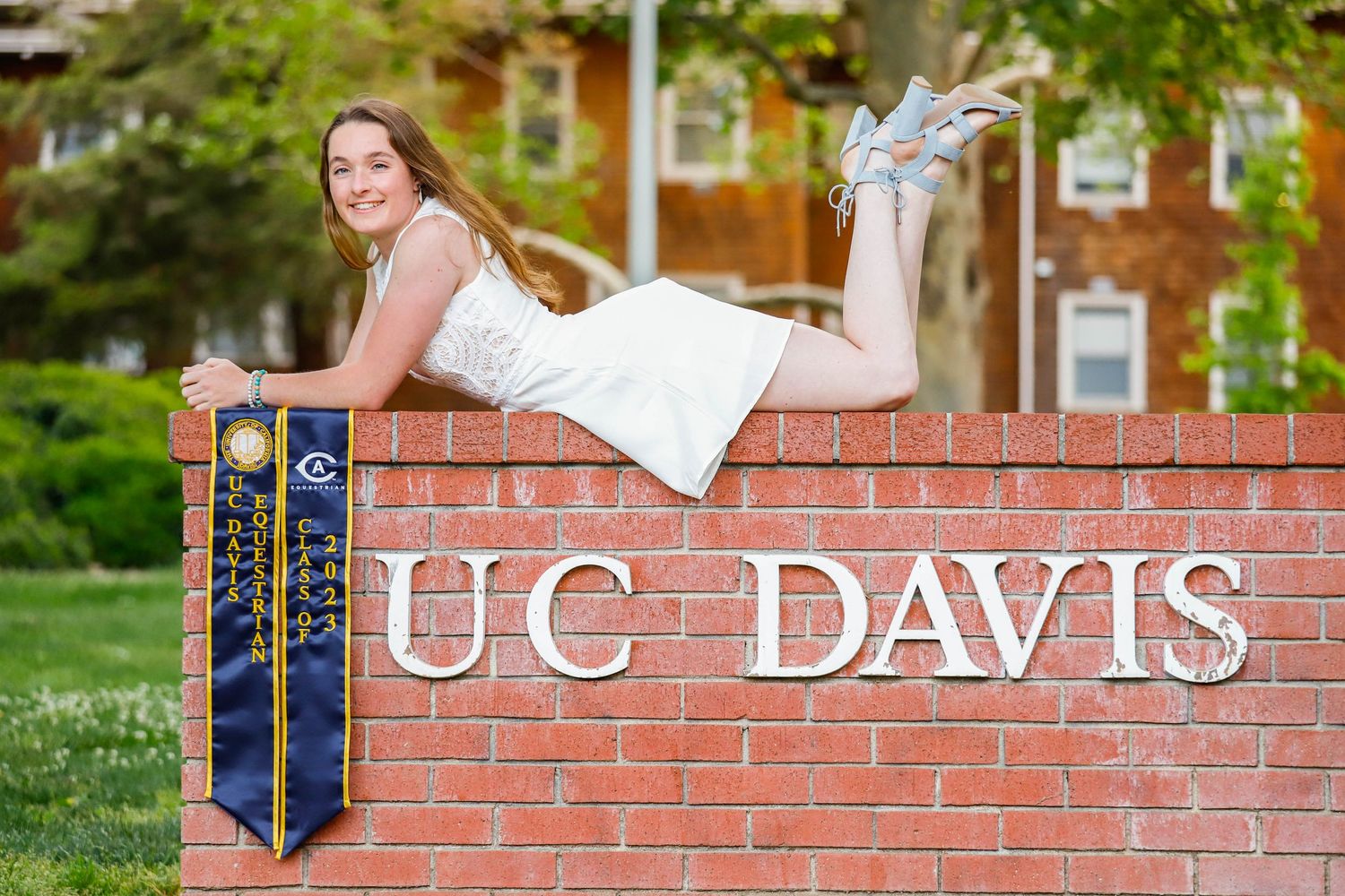 Graduate in white dress posing on UC Davis brick sign with blue and gold stole.