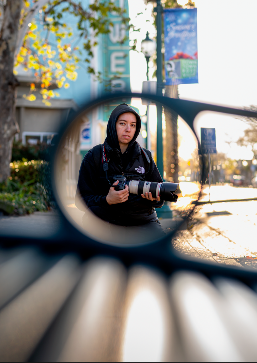 Photographer in black hoodie holds camera with telephoto lens, framed through metal bench circle outdoors.