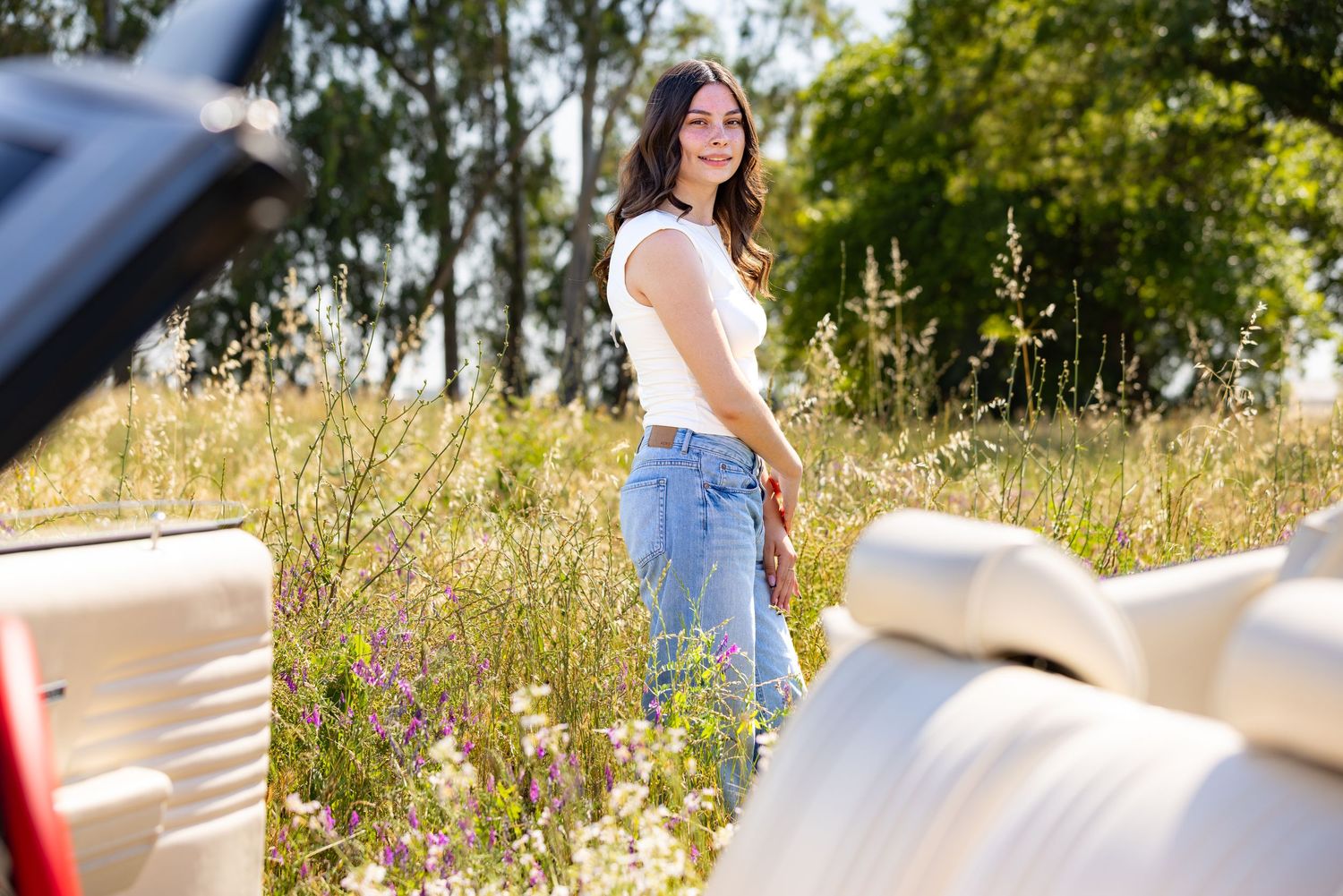 Young woman in white top and jeans smiling in a wildflower meadow beside a vintage convertible car on a sunny day.