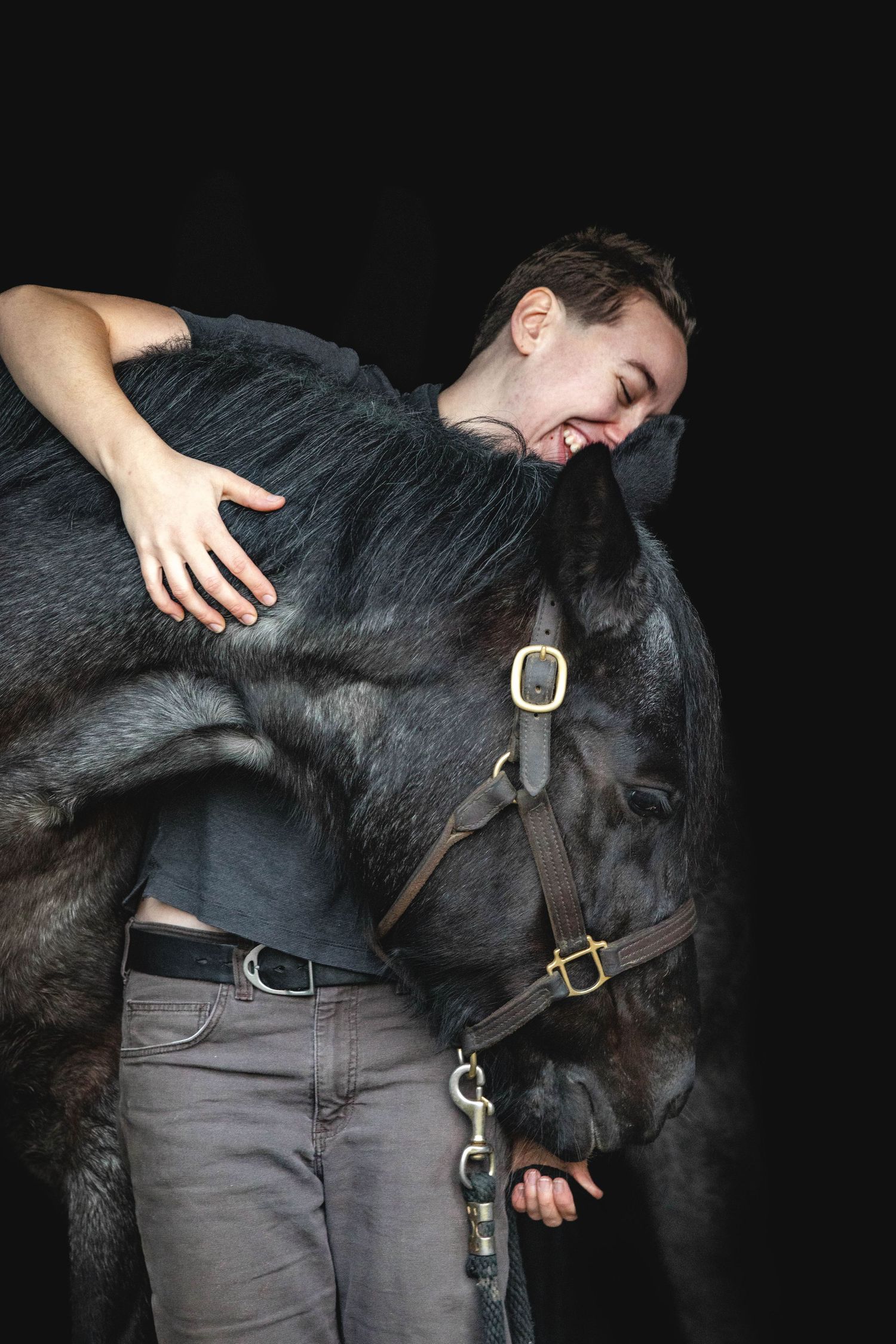 Young man laughing while embracing a dark horse against a black studio background.